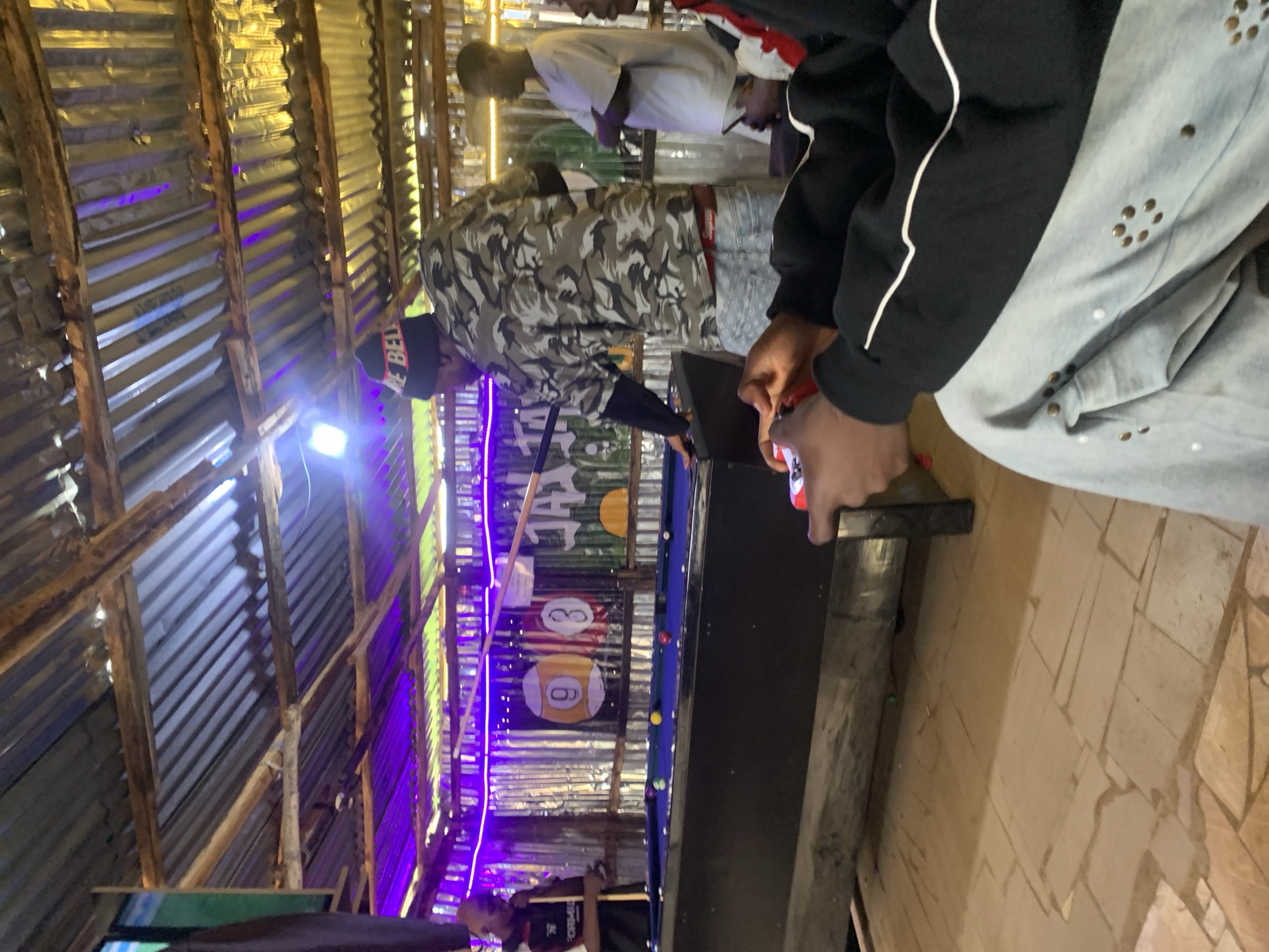 In Durumi, Nigeria, a group of young men are engaged in a game of pool at an outdoor establishment. The scene is dimly lit, with purple and yellow LED lights illuminating the corrugated iron roof and walls. A pool table, set up on a tiled floor, is the central focus. The balls on the table are scattered, with some in the pockets. One man, wearing a camouflage shirt and a black beanie with red lettering, is positioned to strike a ball with a pool cue. Another man, dressed in a black shirt and cap, stands by the table, holding a pool cue. The background reveals more people, some observing the game and others interacting. A screen displaying a green football match is visible on the left side of the frame, suggesting that other entertainment might be available. The overall atmosphere appears casual and social, typical of a local gathering spot.