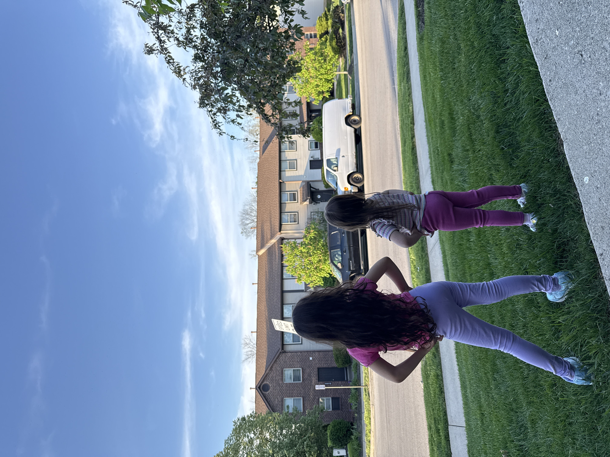 The image captures a residential street scene in bright daylight. Two young girls, viewed from behind, stand on a patch of green grass next to a concrete curb, looking towards the street and houses. The girl in the foreground has long dark hair, wears a pink t-shirt, light purple leggings, and light blue sneakers. Behind her, another girl with dark hair wears a striped pink, white, and purple t-shirt, dark pink leggings, and light-colored sneakers.
The street runs horizontally across the midground, with a white panel van and a partially visible black SUV parked along it. On the opposite side of the street are several two-story houses, varying in architectural style with some featuring brick facades and others siding. Lush green trees are scattered throughout the scene, partially obscuring the houses and reaching into the clear blue sky, which features some scattered, wispy white clouds. A "SPEED LIMIT" sign is partially visible between the two girls, near the head of the closer girl. The overall setting is a sunny, suburban environment in Columbus, United States of America.
