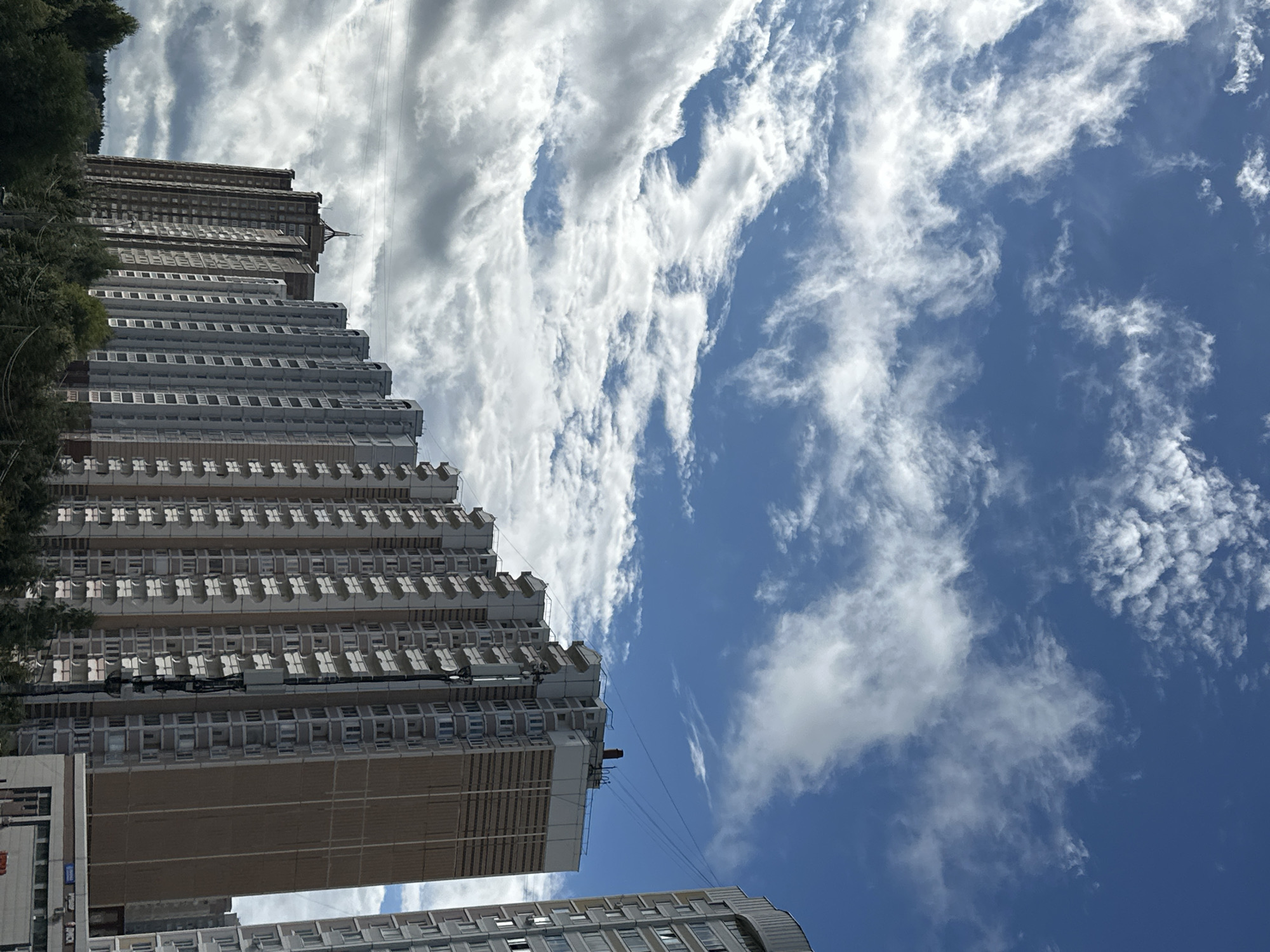The image shows a row of tall residential buildings in the city of Podolsk, Russia, against a backdrop of a blue sky with white clouds. The buildings are modern, with many balconies and windows. The perspective is from ground level, looking up at the structures. Trees and vegetation are visible in the foreground, suggesting a park-like setting or the edge of a residential area. The sky is dynamic, with some areas of bright blue and scattered cumulus clouds, indicating fair weather.