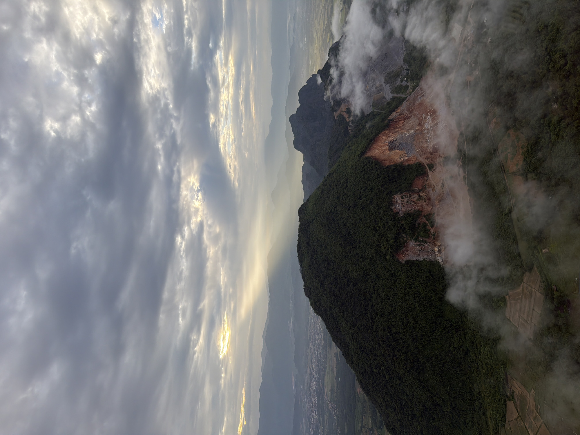 An aerial view captures the rugged landscape surrounding Vang Vieng, Laos, under a dramatic sky. The foreground is dominated by a large, verdant mountain, its slope scarred by a visible mining operation with exposed reddish-brown earth and grey rock. Pockets of white mist cling to the lower slopes and drift into the valley below, obscuring parts of the landscape.
In the mid-ground, jagged, dark mountain peaks rise, also partially shrouded in mist. A dusty, winding road is visible on the slopes of these mountains, likely leading to the quarry. Beyond these mountains, the town of Vang Vieng spreads out, a cluster of buildings and fields, with more distant, hazy mountain ranges forming the backdrop. The sky is filled with heavy, grey clouds, but shafts of sunlight pierce through, illuminating patches of the scene and creating a sense of depth and atmosphere. The overall impression is one of natural beauty juxtaposed with industrial activity.