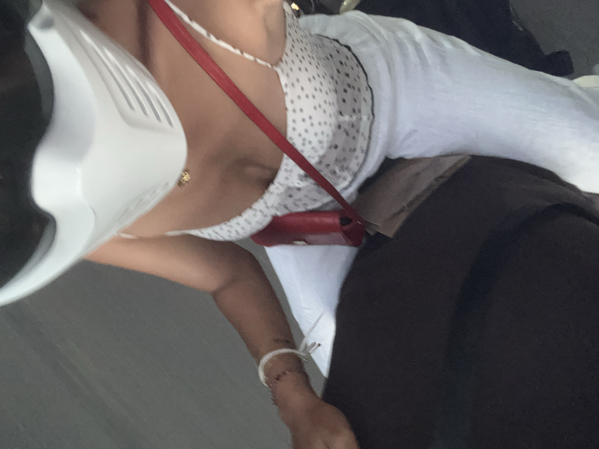 A person wearing a white motorcycle helmet, a white polka-dot tank top, and white pants rides a motorcycle. A red purse is slung across their chest. The person has a bracelet on their left wrist. The background is a blurred grey surface, consistent with a road. The scene is captured from a first-person perspective, likely from another motorcycle.