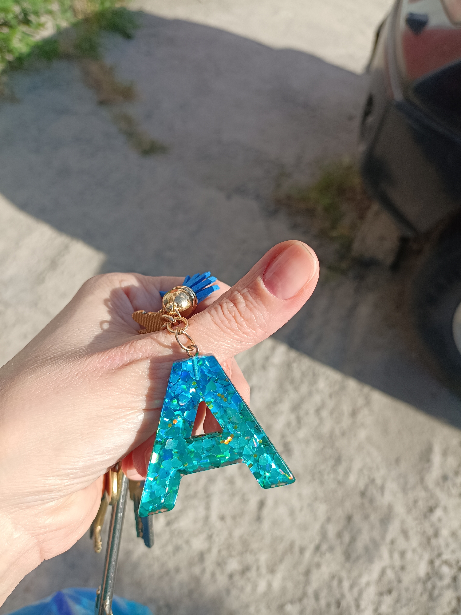 A close-up shot shows a person's thumb and fingers holding a keychain with the letter "A" made of turquoise resin with glitter. The keychain also has a small gold charm shaped like a dog and a blue tassel. Several keys are attached to the keychain. The background is a concrete surface with shadows, and a blurry black car is visible on the right side. This scene appears to be outdoors, possibly in a parking lot, given the concrete and the car. The lighting suggests it is daytime. The image does not provide any specific information about the location within the city of Bataysk, Russia.