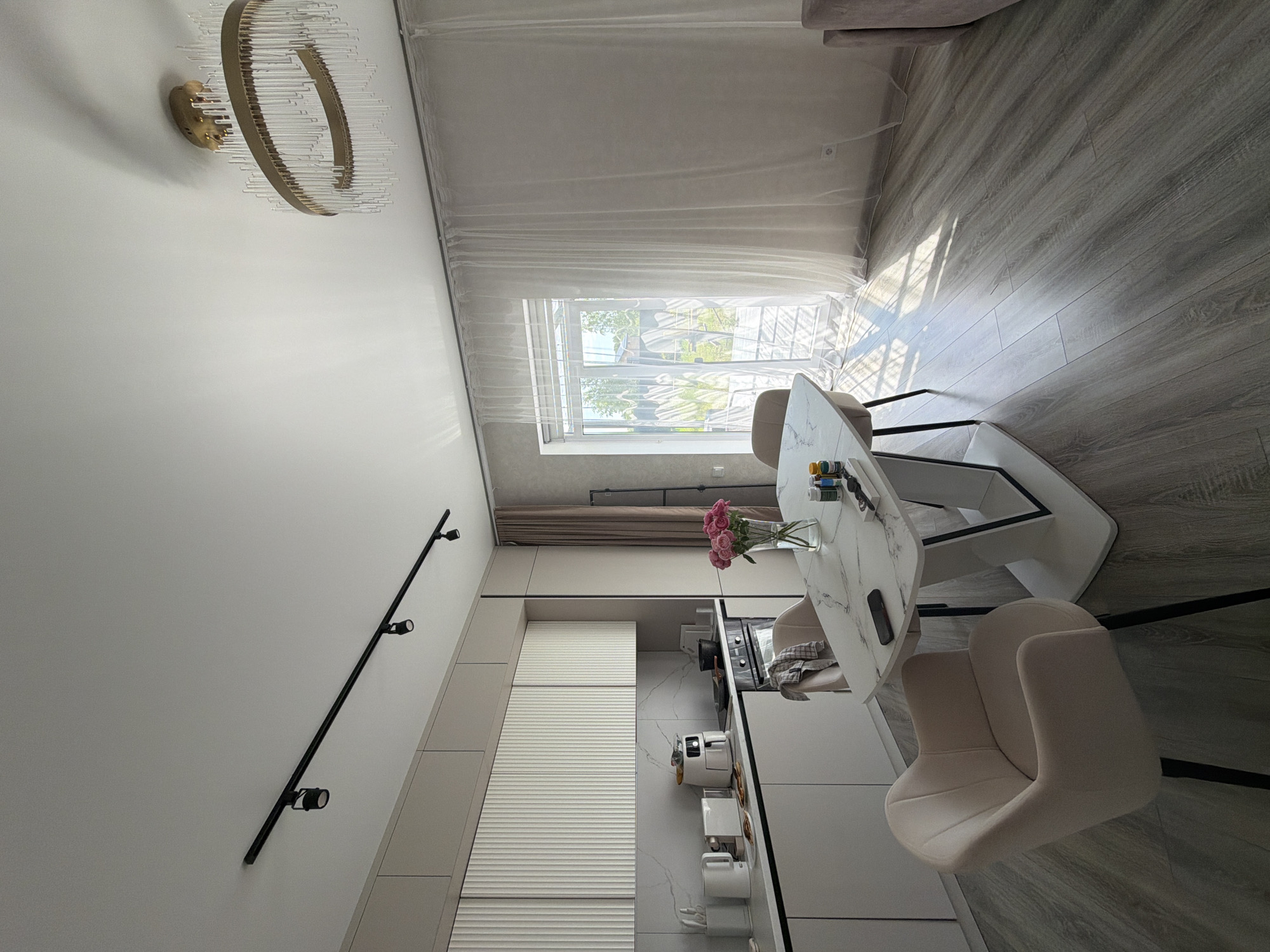 In Malinovka, Russia, a modern kitchen and dining area is depicted. The scene is brightly lit by natural sunlight streaming through sheer white curtains covering a large window or glass door. The room features light-colored cabinetry with fluted detail on the upper cabinets. A marble-topped dining table with a white pedestal base is positioned in the center of the room, surrounded by two upholstered armchairs in a pale blush color. On the table, a vase of pink roses, a smartphone, and several small bottles of what appear to be art supplies or medicines are visible. The kitchen counter has two white appliances, possibly a coffee maker and a toaster, along with some plates. A black oven is integrated into the cabinetry. The floor is covered with grey wood-look laminate flooring. A circular chandelier with glass rod accents hangs from the ceiling, and a black track lighting fixture is also present. The overall impression is one of a clean, contemporary, and well-lit living space.