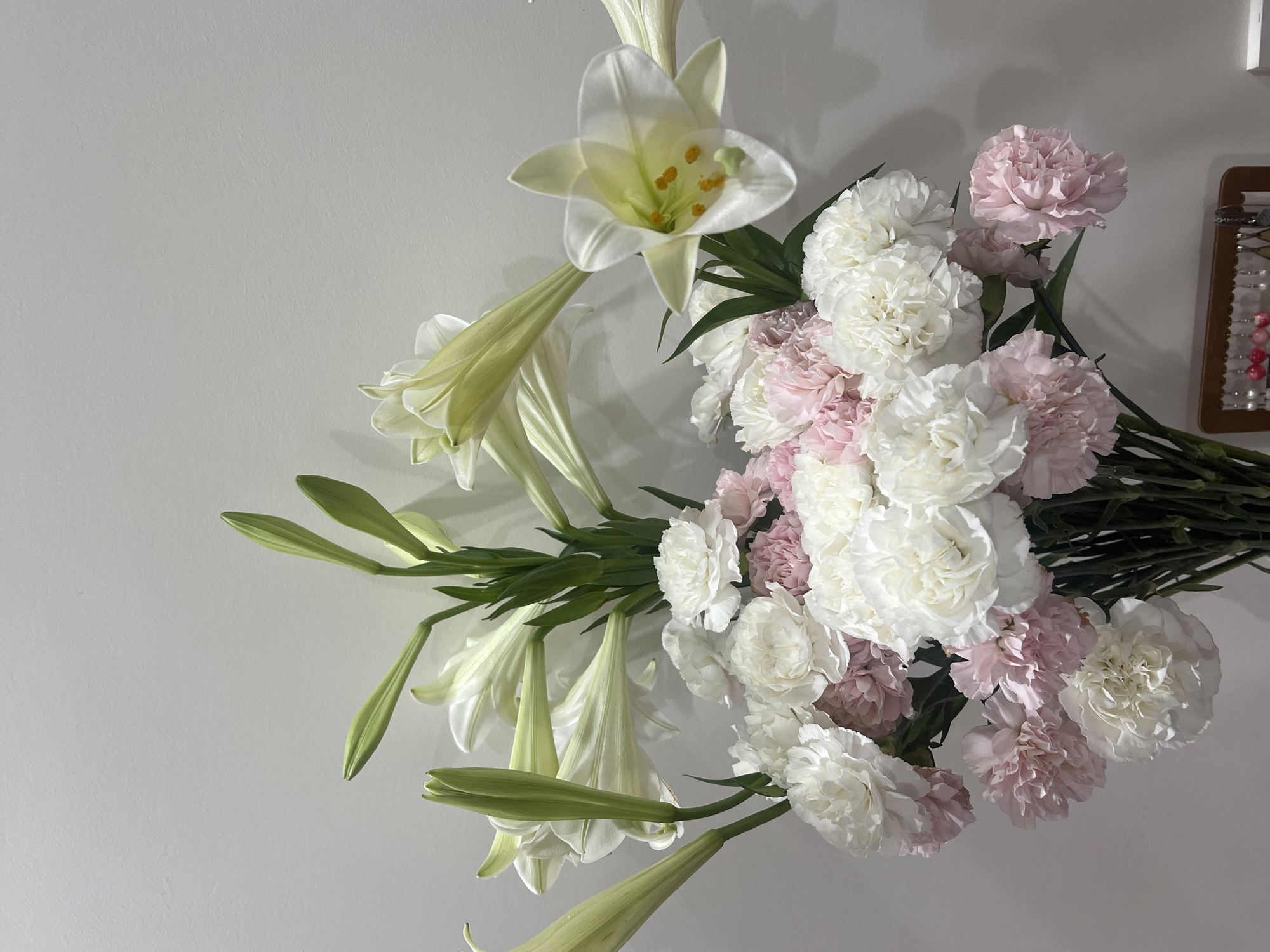 A high-angle shot depicts a bouquet of white lilies and pink and white carnations against a plain, light grey background. The lilies are mostly unopened buds except for one fully opened bloom. The carnations are densely clustered. A small decorative item with beads is partially visible in the upper right corner. The setting appears to be indoors, with neutral lighting. The city of Nam Nha Trang Ward, Vietnam, is not visibly represented in the image.