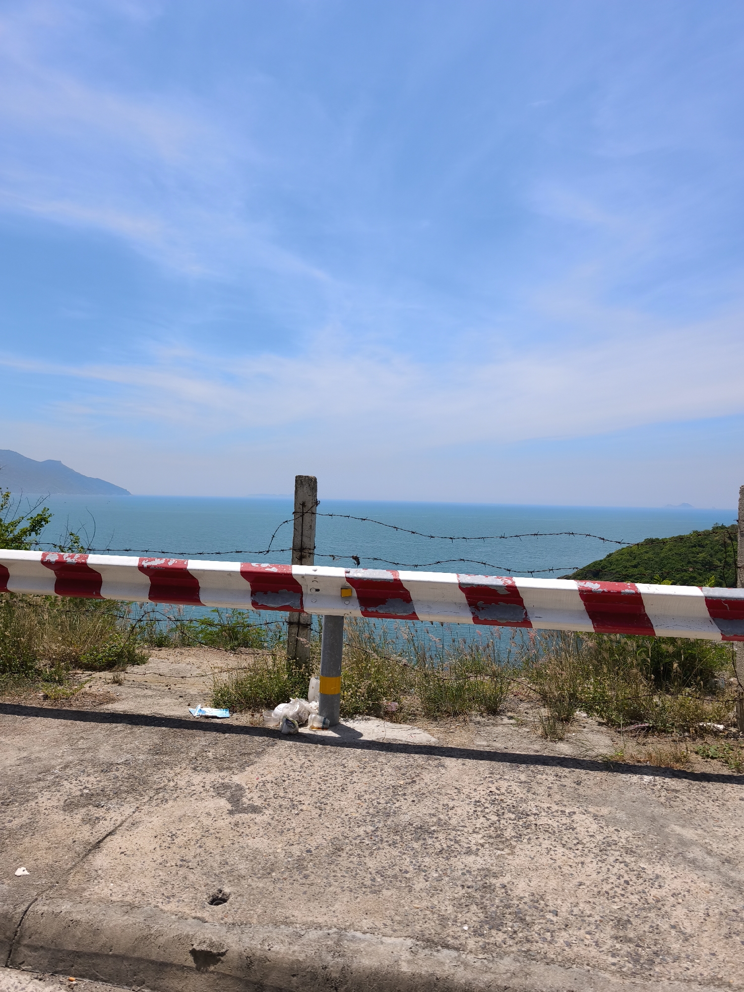 The image captures a scenic overlook in Bac Nha Trang Ward, Vietnam, featuring a striking view of the turquoise ocean under a clear blue sky dotted with wispy clouds. In the foreground, a weathered guardrail with red and white stripes runs horizontally across the frame, partially obscured by barbed wire. Concrete poles support the guardrail, one of which is positioned on the right side of the image. The ground in front of the guardrail is a rough, concrete surface, showing signs of wear and tear, with scattered patches of dry grass and some litter, including plastic bags and cups, near the base of the pole. Beyond the guardrail and barbed wire, the vast expanse of the ocean stretches towards the horizon, with distant hills and islands visible in the background, their outlines softened by the atmospheric haze. The overall impression is one of natural beauty contrasted with the subtle presence of human infrastructure and the unfortunate evidence of litter.