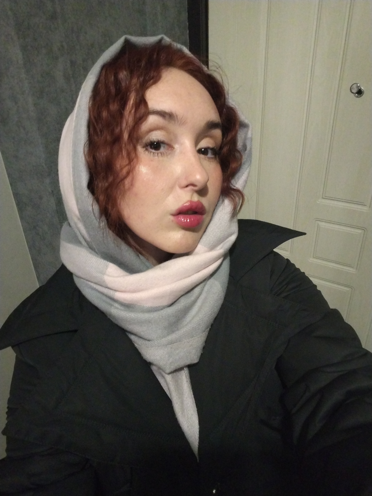 A woman with fair skin and auburn, curly hair, partially covered by a light gray and pink patterned headscarf, poses for a close-up photograph. She has a black jacket with a wide collar. The background includes a gray patterned wall to the left and a white door with vertical panels and a round doorknob on the right. The location is not discernible as Izhevsk, Russia, from the visible elements in the image.