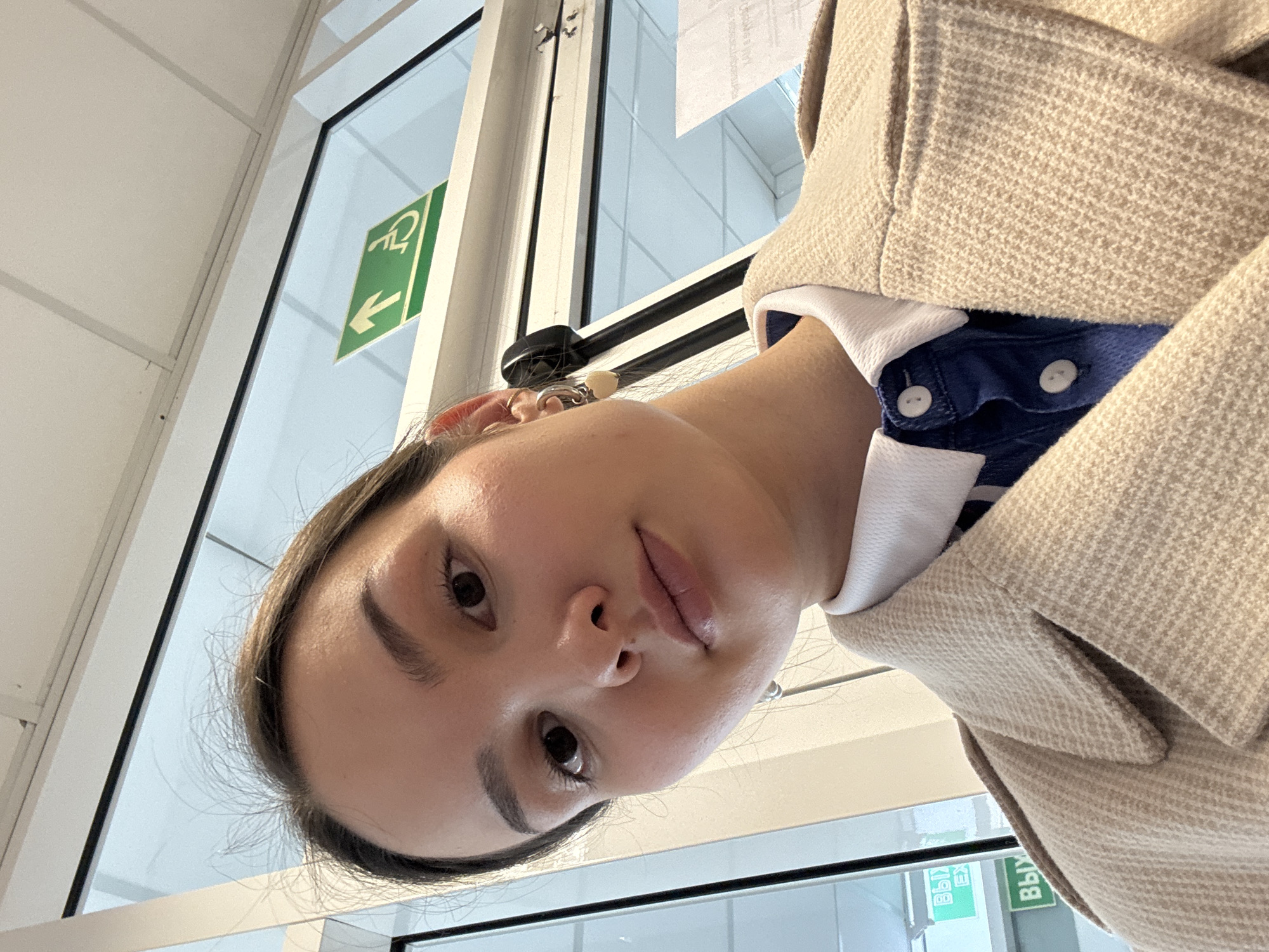 A young woman is taking a selfie indoors, in what appears to be a public building in Chaikovsky, Russia. The selfie is taken from a low angle. The woman has brown eyes, dark eyebrows, and light skin. She is wearing a blue shirt with white buttons, a white undershirt, and a light brown wool jacket. She has a silver earring in her ear. In the background, there is a glass door with a green sign that features a wheelchair symbol and an arrow. There are also some signs in the Russian language visible on the door.