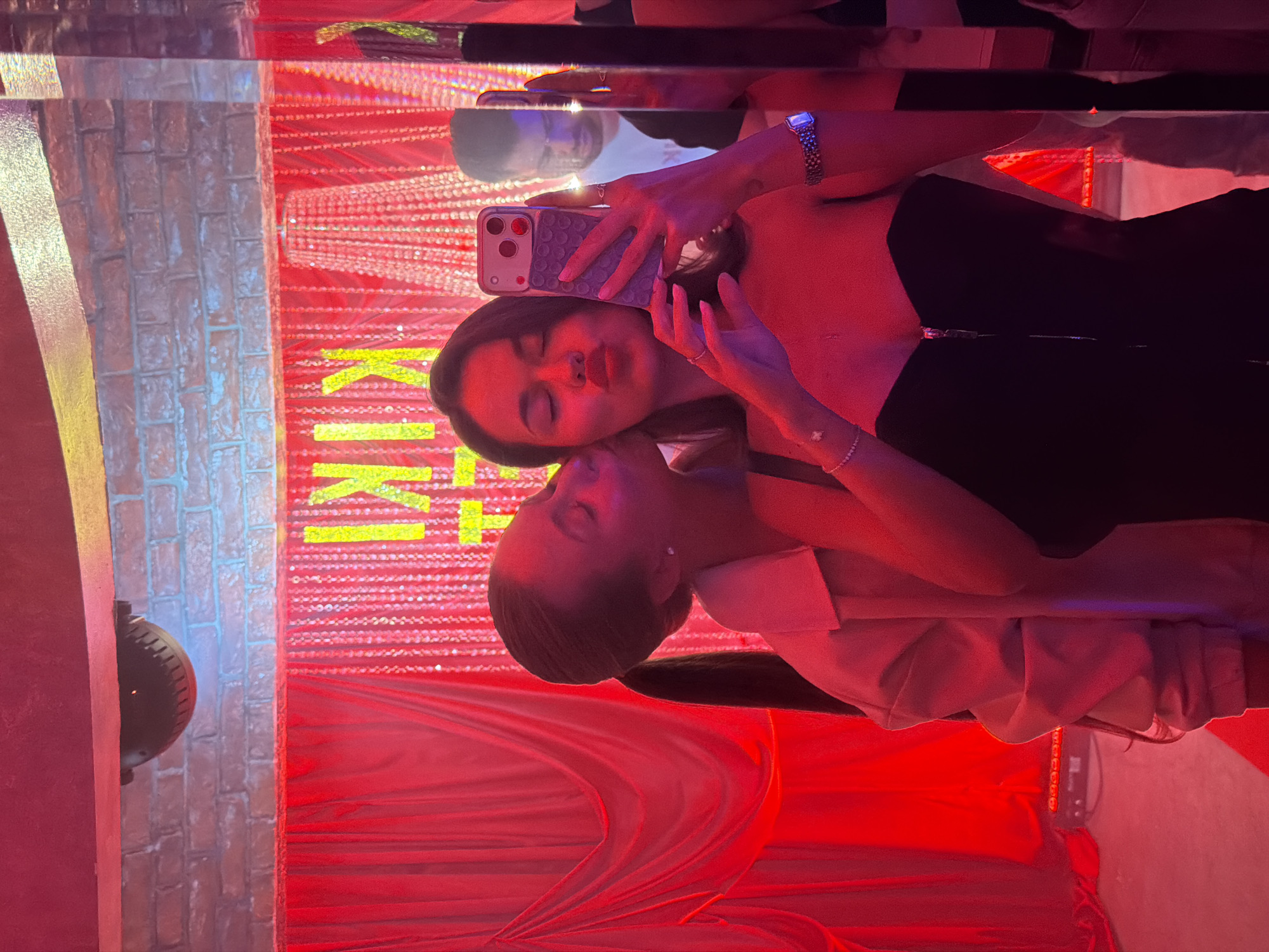The image captures a mirror selfie featuring two females and a male reflection within an entertainment venue, KIKI bar/club, located in Moscow, Russia. The scene is illuminated predominantly by intense red artificial light.
In the foreground, a female with dark hair is holding a light-colored smartphone with a three-lens camera in her right hand, taking the photograph. She has puckered lips and closed eyes, wearing a black strapless top with a front zipper, a bracelet with a cross-shaped charm on her left wrist, and a silver-toned rectangular watch on her right wrist. To her left, a second female, also with dark hair tied in a ponytail, is kissing her cheek. This individual wears a light-colored collared garment, and her eyes are closed. In the reflection visible to the right, a male individual with short dark hair, wearing a white T-shirt, is looking downwards or to his left.
The background features a brick wall spanning the upper portion, beneath which are red fabric drapes on the left and a shimmering, beaded curtain on the right. Behind the beaded curtain, the name "KIKI" is illuminated vertically in yellow script. A dark, dome-shaped device, possibly a security camera or light, is mounted on the ceiling above the brick wall. Subtle blue lighting is visible along the top of the brick wall on the left. The floor in the lower left is covered in red material, with dark equipment visible on its surface.