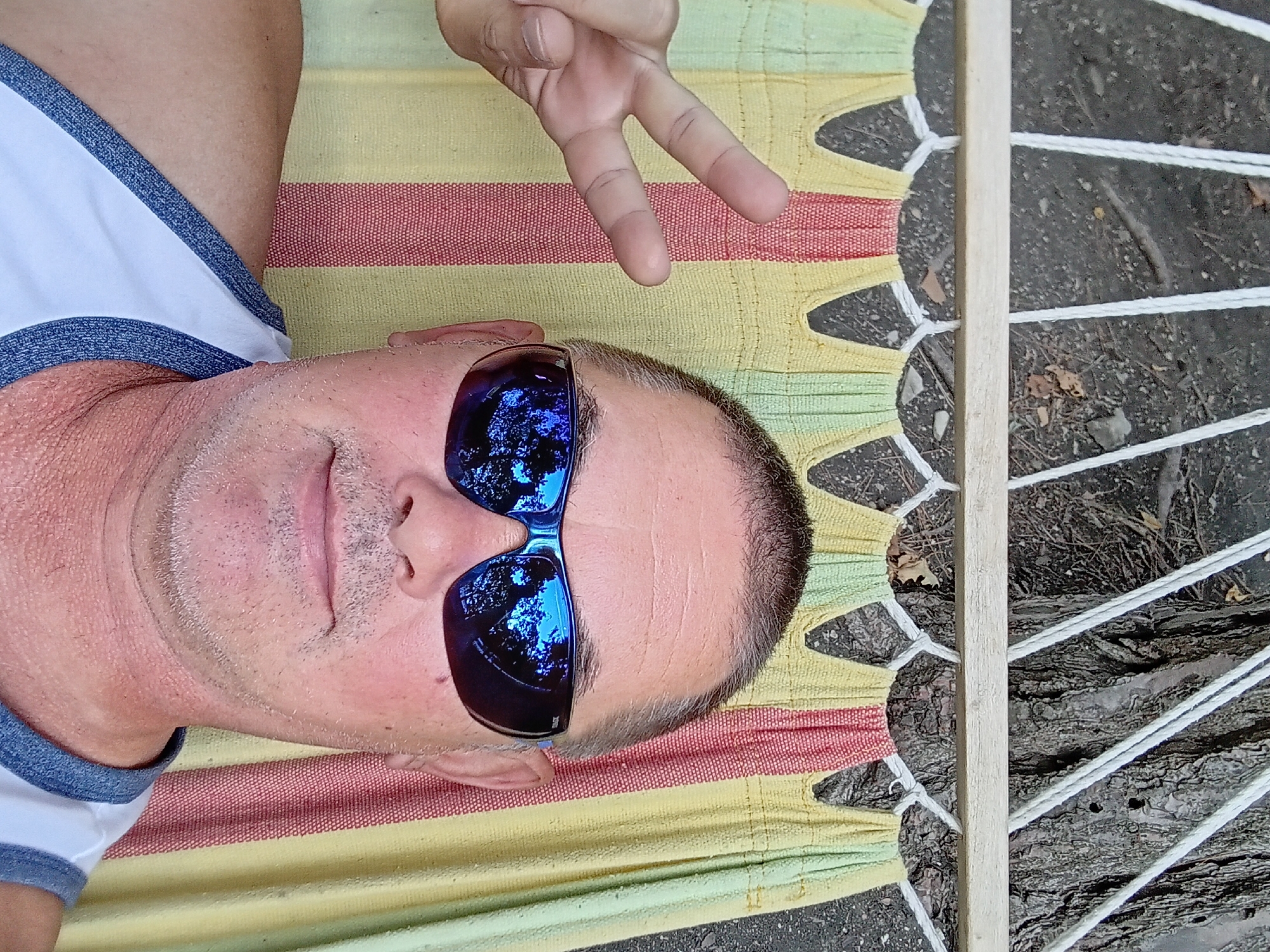 A man with sunglasses is lying in a hammock. He is wearing a white tank top with blue trim. He is holding up two fingers in a peace sign. The hammock has yellow, red, and green stripes. In the background, there is dirt and a tree.