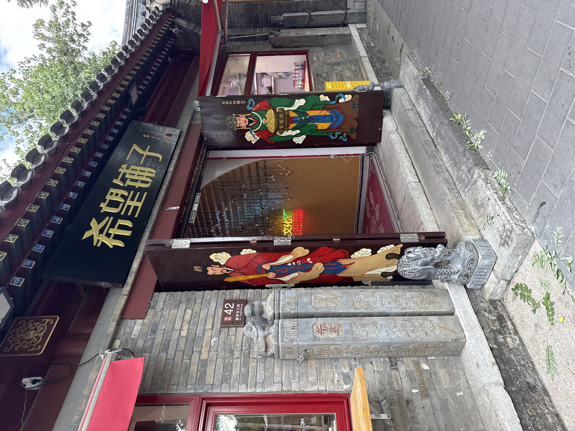 The image depicts the exterior of a shop on St. Yonghegong Street in Dongcheng District, China. The shop features a traditional architectural style with grey brick walls, a dark red roof with decorative tiles, and dark brown doors. The shop's name, "希望铺子" (Xiwang Puzi), is displayed in golden Chinese characters above the entrance. The doors are adorned with painted figures, one on each door, depicting what appear to be traditional deities or historical figures in colorful attire.
In front of the shop, there are two stone pillars with carved designs and what look like small stone lion or dog statues on top. A sign with the number "42" and Chinese characters "雍和宫大街" (Yonghegong Dajie) is mounted on the wall to the left of the entrance, indicating the street address. A red awning is visible on the left side of the building. Inside the shop, visible through the open doors, there are hanging decorations and a neon sign that reads "文化" (Wenhua), meaning "culture".
The ground in front of the shop is paved with grey bricks, and there are some fallen leaves scattered on the ground and along the edge of the sidewalk. The environment appears to be an urban street with a mix of traditional and modern elements, given the architecture and the presence of the neon sign. The overall impression is of an established business in a historic part of the city.