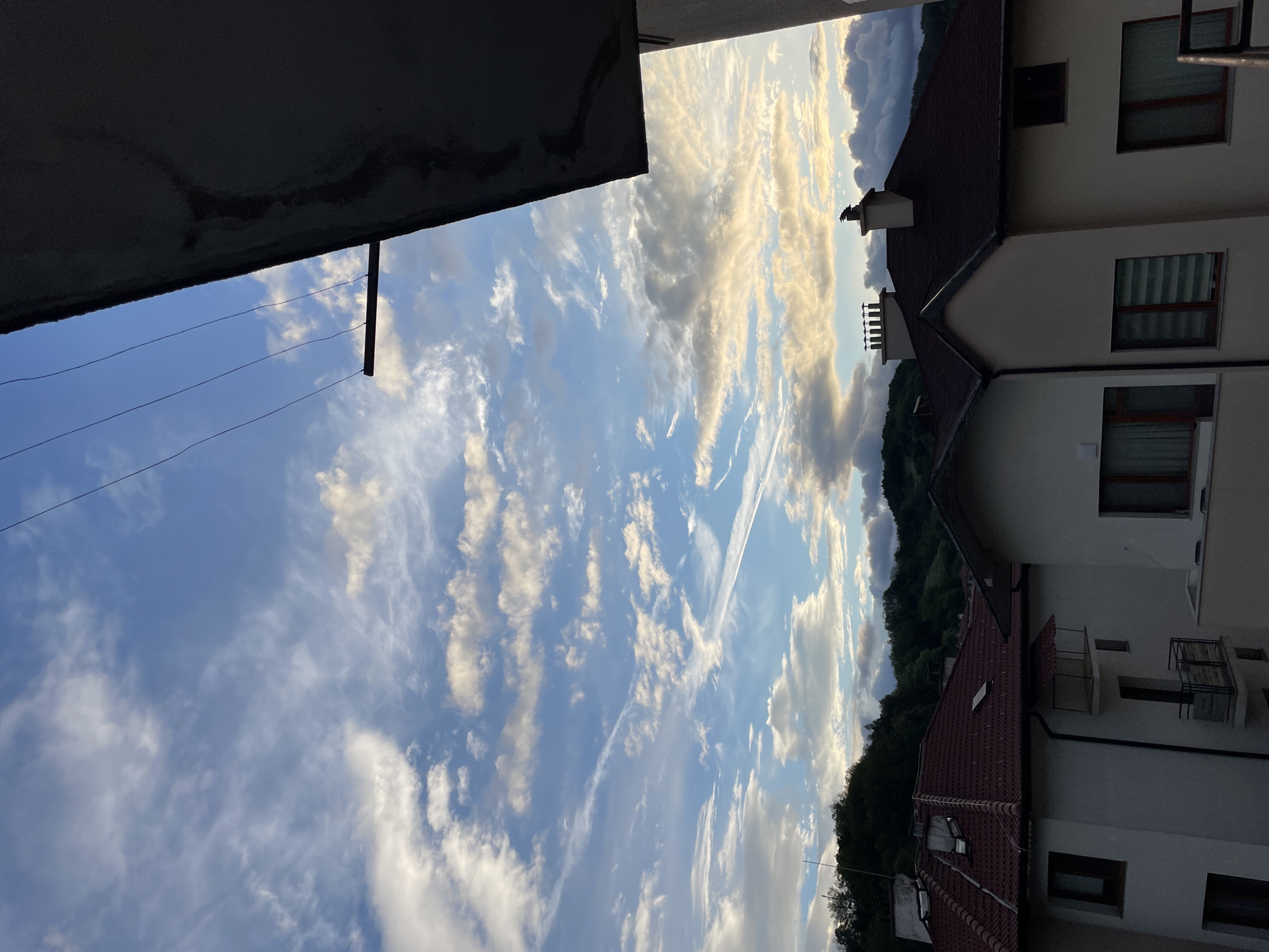 The image depicts a view in Zlatograd, Bulgaria, looking upwards at a partially cloudy sky. The sky is blue with streaks of white clouds, and there are visible contrails from aircraft. In the foreground on the left is a section of a building, likely a balcony or part of a concrete structure, obscuring some of the sky. Two power lines are visible against the sky. On the right side of the frame, there is a view of adjacent buildings with red tiled roofs and white facades. Forested hills are seen in the background. The composition is somewhat tilted.