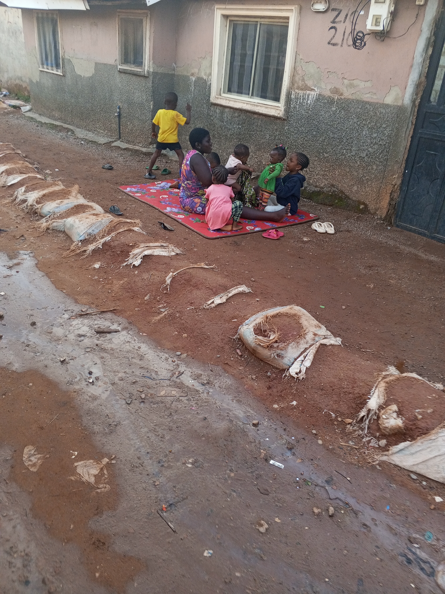 The image depicts a scene on a red dirt road in what appears to be a low-income neighborhood. A woman sits on a red patterned mat on the ground outside a building. She is surrounded by four children. One child, wearing a yellow shirt, stands beside the building. The other children are near the woman, with one in her lap. The ground near the road is muddy and littered with debris. Sandbags are positioned along the side of the road, possibly as erosion control. The building has a decaying exterior with two windows on the left and one on the right of the image. Wires and some numbering are painted on the building facade near the top right. A partially visible dark doorway is present to the far right. The city and country are provided by the prompt, but do not appear visually in the scene.