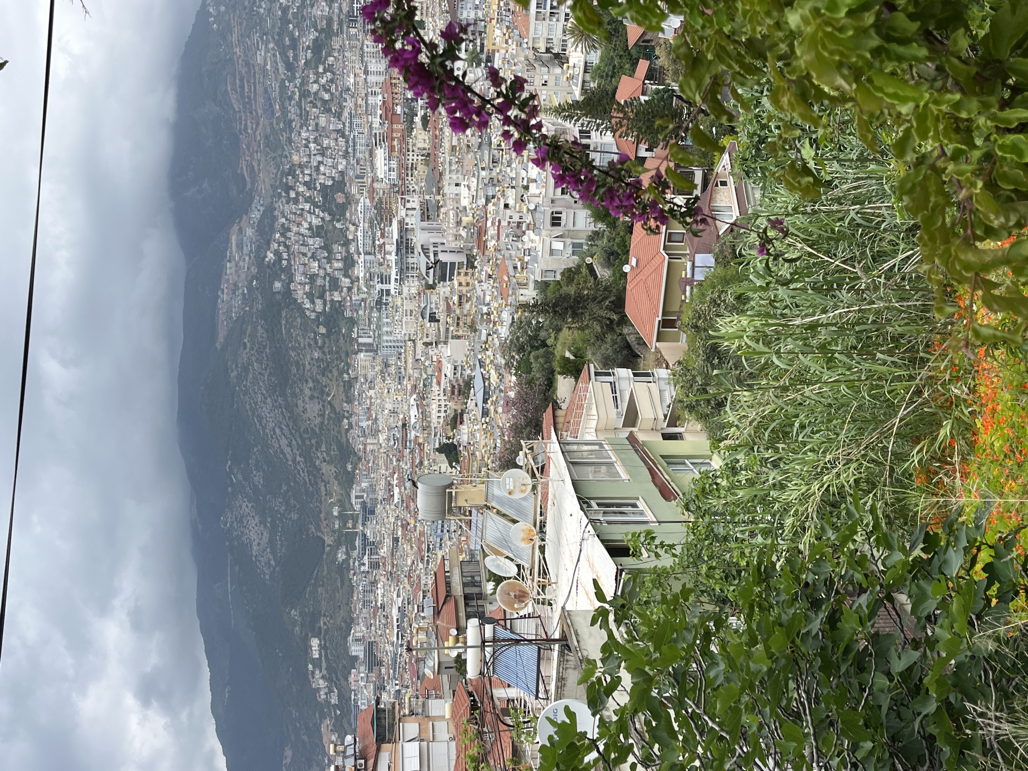 The image is an elevated view of Alanya, Turkey. In the foreground, there are buildings with satellite dishes and water tanks on the rooftops, interspersed with dense greenery and flowers. The middle ground presents a cityscape with tightly packed buildings of varying heights and colors, giving a sense of urban density. In the background, a large mountain range is partially obscured by heavy cloud cover. The overall atmosphere is slightly overcast, with the light subdued by the cloud cover.