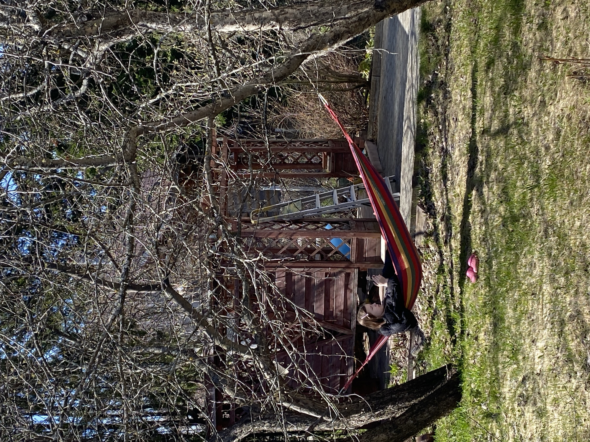 A person is lying in a multicolored hammock outdoors. The hammock is strung between two trees. A small wooden building is in the background behind the trees. There is a ladder leaning against the building. A pair of pink shoes is on the ground near the hammock. The ground is covered in grass and fallen leaves. The trees are mostly bare. The location may be Прикамский, Russia.
