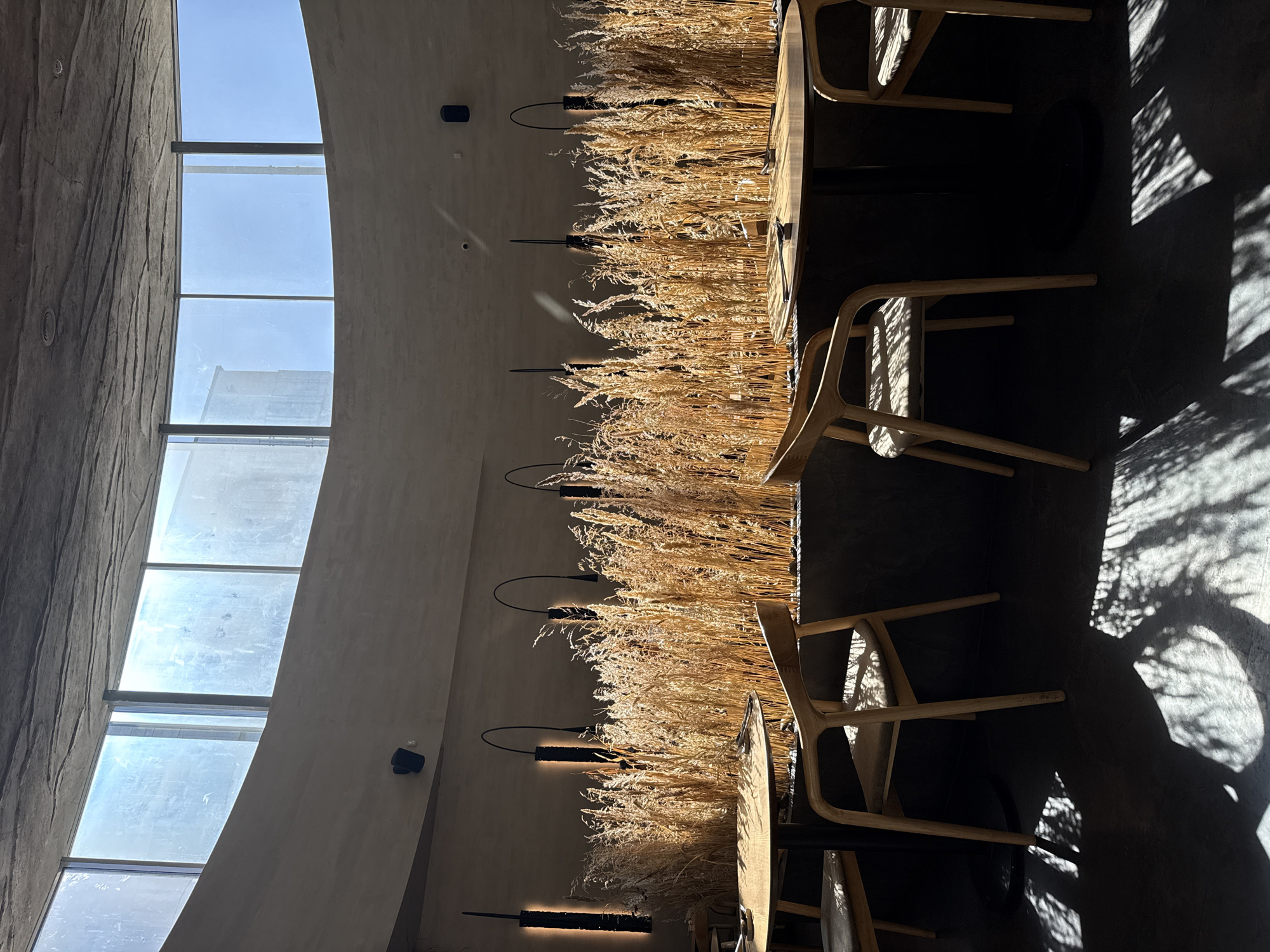 The interior of a restaurant in Krasnoyarsk, Russia, features a row of dining tables with wooden chairs adjacent to a curved wall with a large window. The tables are decorated with tall, dry grass arrangements. Above the grass are mounted black, industrial-style sconces. Light streams through the windows, creating shadows on the floor.
