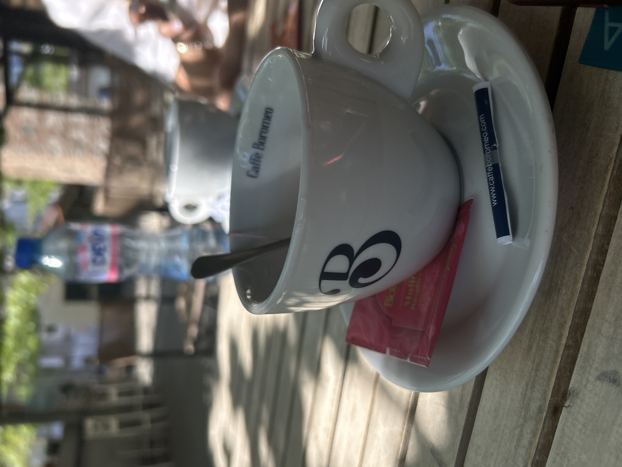 The image shows a table with two cups of coffee and a bottle of water. One cup is on a saucer with a sugar packet and a stirring stick, and has the brand "Caffe Borromeo" printed on it. The table is made of wood and appears to be located outdoors, given the presence of dappled sunlight and trees in the blurred background. While the exact location cannot be forensically determined from this image, based on the subject matter (coffee, outdoor setting), it's plausible that the picture was taken at a cafe or restaurant in Sofia, Bulgaria.