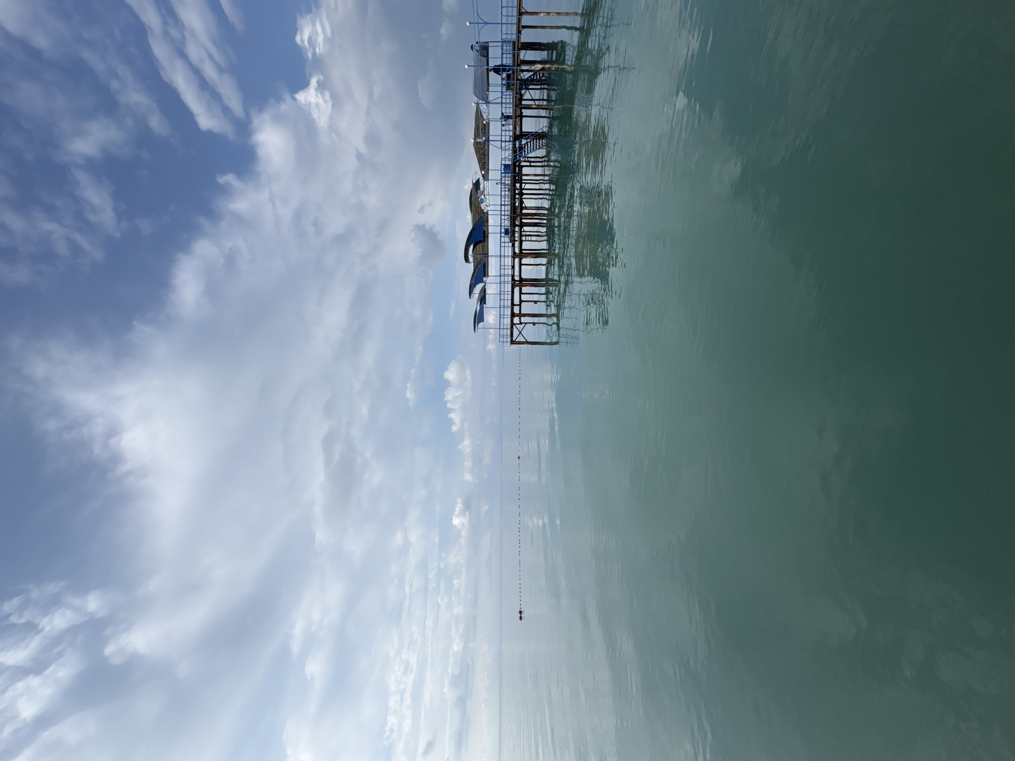 This image captures a serene view of a pier extending into a calm, turquoise body of water, likely Issyk-Kul Lake in Sary-Oy, Kyrgyzstan. The sky above is a mix of blue and white clouds, with wispy formations scattered across the expanse. The pier, constructed of wood and metal, features a blue railing and several covered seating areas, suggesting a recreational or bathing area. A person is visible on the pier, standing near the railing. The water is placid, reflecting the sky and pier, with a line of buoys or markers creating a subtle barrier in the distance. The overall atmosphere is peaceful and tranquil, evoking a sense of calm before or after a day of activity.