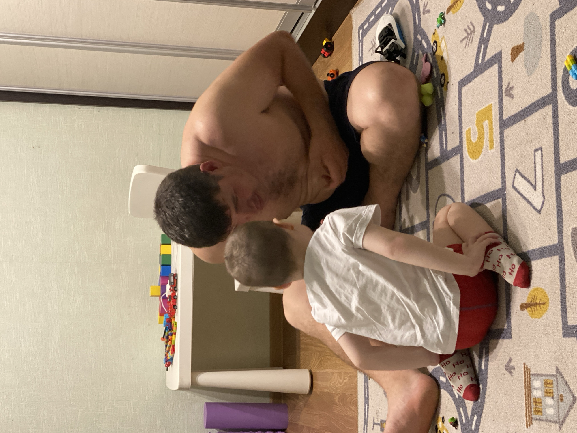 A man is kneeling with his son on a patterned rug. The man is shirtless and wearing dark shorts. The boy is wearing a white t-shirt, red shorts, and "Ho Ho" patterned socks. A white table is to the left, adorned with toys. The rug is multi-colored with patterns resembling a hopscotch game. Other toys are scattered around. A wall is behind the table, and a sliding closet door is visible above the man. The setting is a domestic indoor environment. The city of Sochi, Russia is not explicitly indicated in the image.