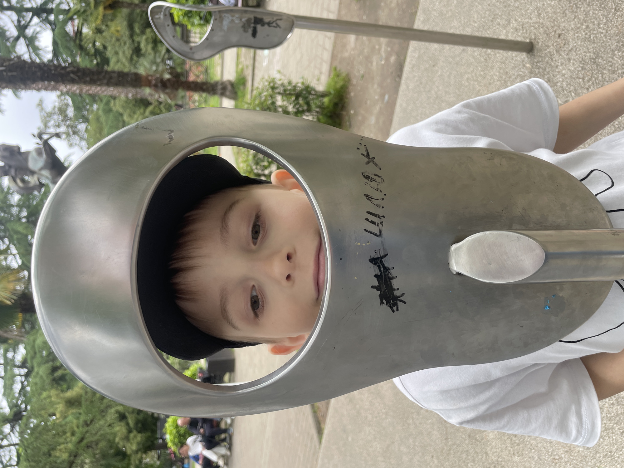 A young boy wearing a black cap and a white t-shirt is peering through a metal sculpture. The sculpture features a circular opening through which the boy's face is visible, and it is mounted on a metal pole. The scene appears to be outdoors in a park or public space. In the background, greenery including trees and bushes are visible, along with the blurry figures of people and other similar sculptures. The location is Sochi, Russia.