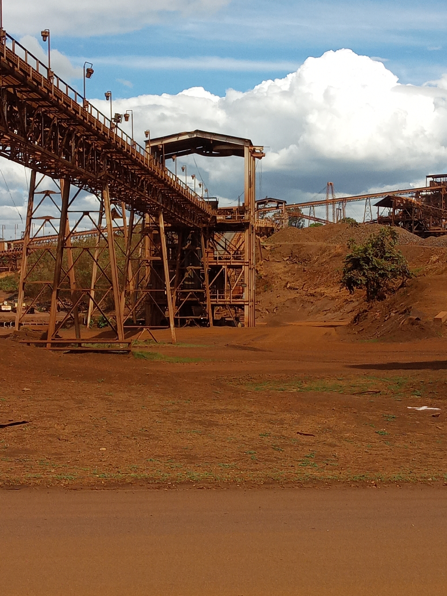 The image depicts an industrial site in Guayana City, Venezuela, characterized by rust-colored earth and oxidized metal structures. Dominating the scene are elevated conveyor systems supported by steel frameworks. The sky is partly cloudy with white cumulus formations. The ground is barren with sparse vegetation. There are no people visible. The site appears to be a mining or processing facility.