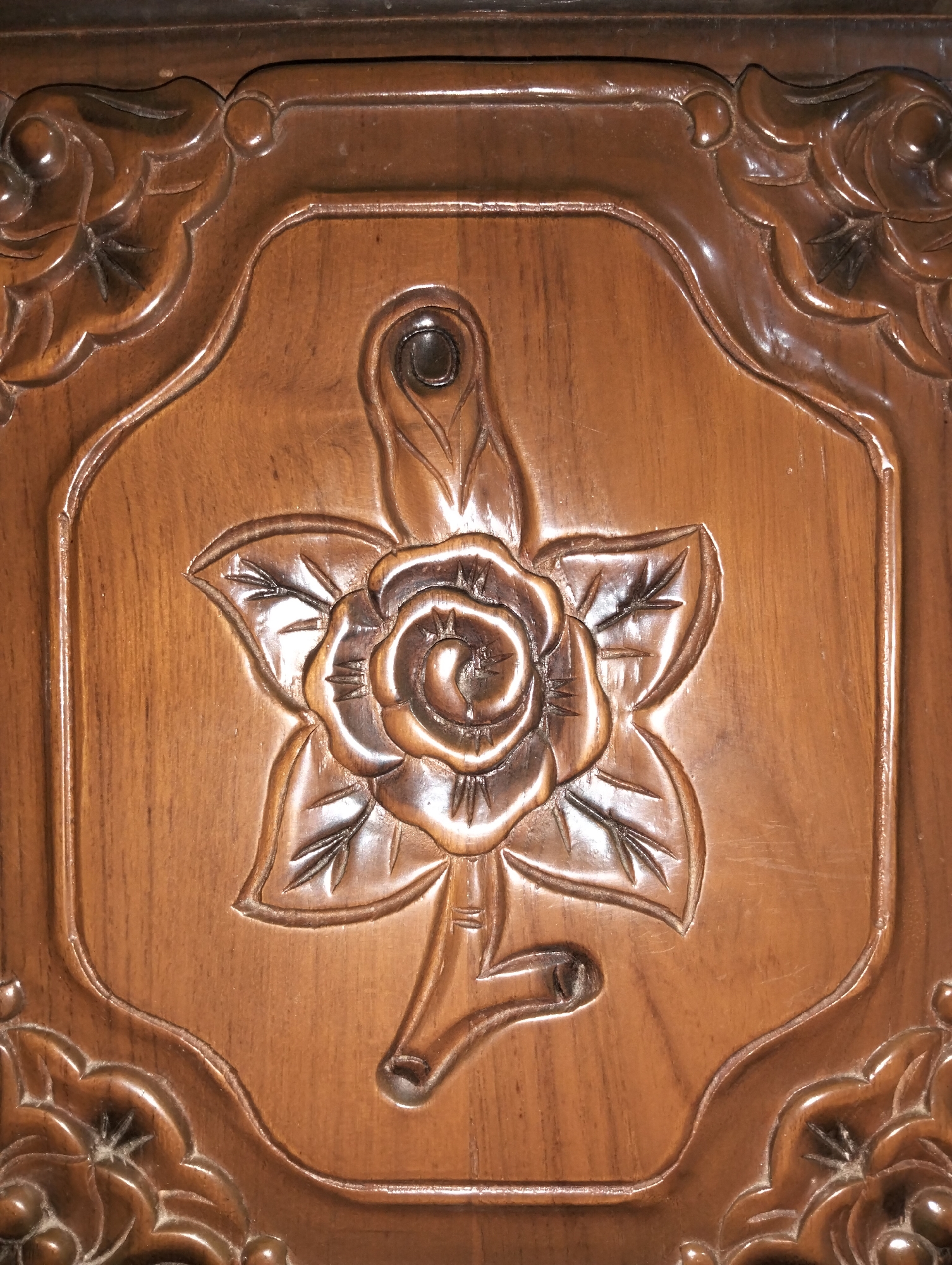 The image shows a close-up of a carved wooden surface. The carving depicts a rose with five petals and a stem with leaves. The wood is a medium brown color and appears to be lacquered. The carving is detailed, with individual petals and leaves clearly defined. The background is a flat, smooth surface of the same wood.
There is no information provided about the location in the city of Payagaboh, Indonesia, and therefore it cannot be included in the description.