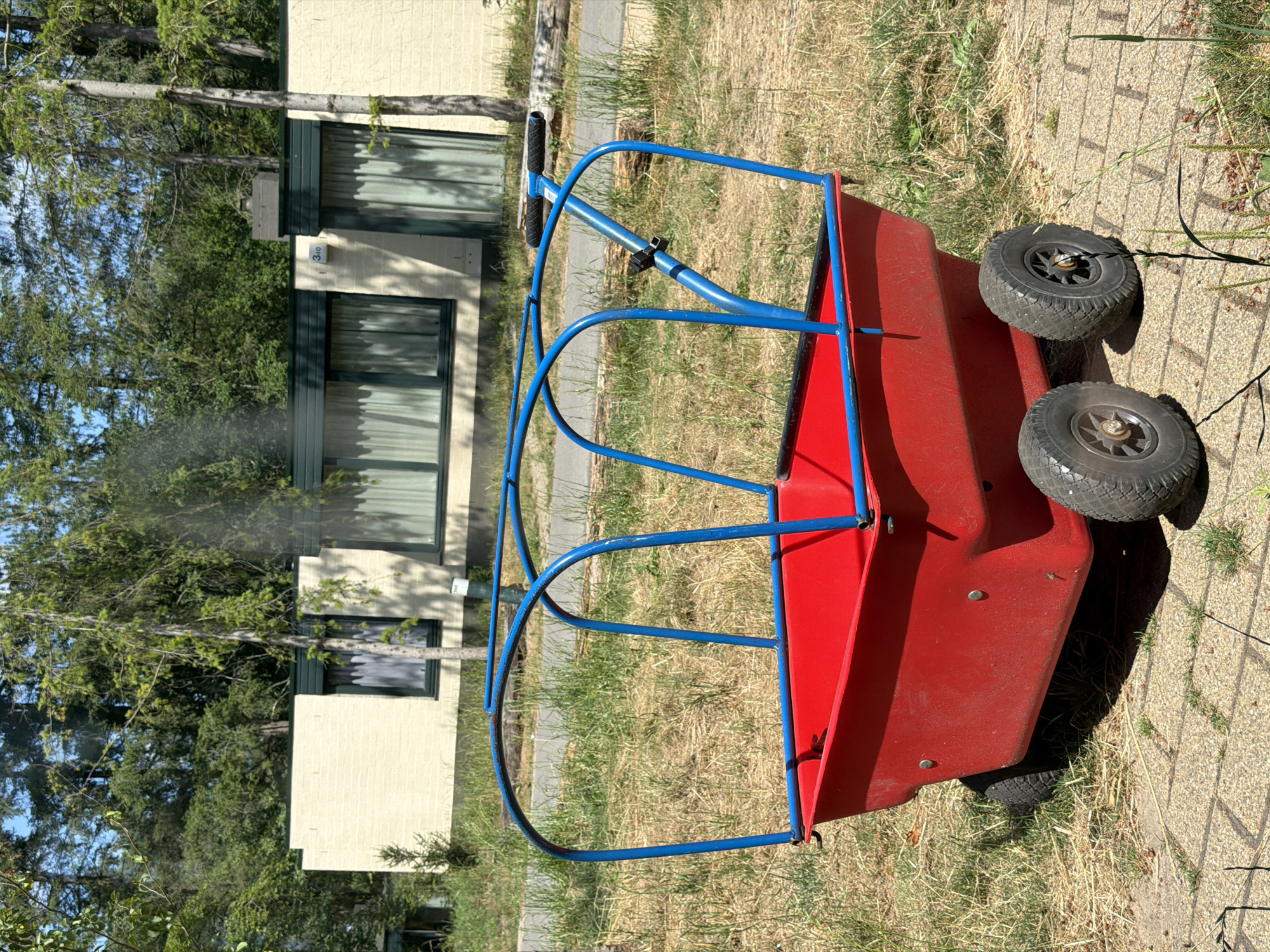A bright red wagon with a blue frame stands on a paved surface in front of a low, cream-colored building with green trim and windows. The building is surrounded by dry grass and trees, suggesting a park-like or recreational setting in Heijen, Netherlands. The wagon has two large wheels in the back and appears to be unoccupied. The sky is clear and blue, indicating a sunny day.