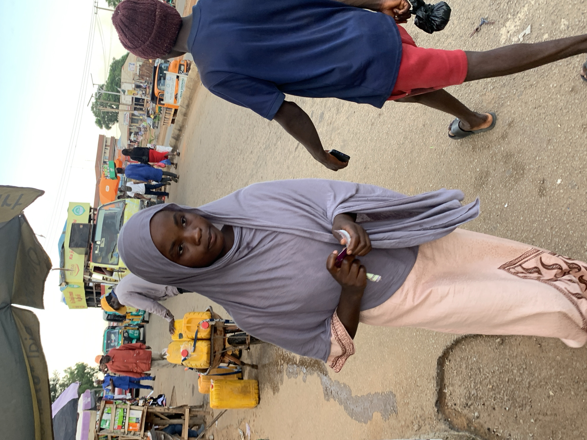 This is a full shot, taken at eye level, of a street scene in Jos, Nigeria, depicting a busy marketplace environment. In the foreground, a young Black female, wearing a light purple hijab and a pink skirt with patterned trim, is walking towards the viewer, holding what appears to be a small item in her right hand. To her right and slightly behind her, another Black individual, wearing a navy blue t-shirt and red shorts, walks away from the camera, holding a black plastic bag in their right hand.
The background reveals a bustling street with various elements of commerce. Several yellow jerrycans, likely containing liquids such as water or fuel, are piled on a cart to the left of the central figures. A light-colored truck with green and yellow signage is visible further down the street, suggesting commercial activity. Other individuals are seen in the middle ground, some appearing to be vendors or shoppers, with stalls and umbrellas indicating market stalls. The ground is paved with dirt and asphalt, with some patches of wetness and debris. The sky is overcast, suggesting diffused daylight. The overall impression is a snapshot of daily life and economic activity in a Nigerian urban setting.