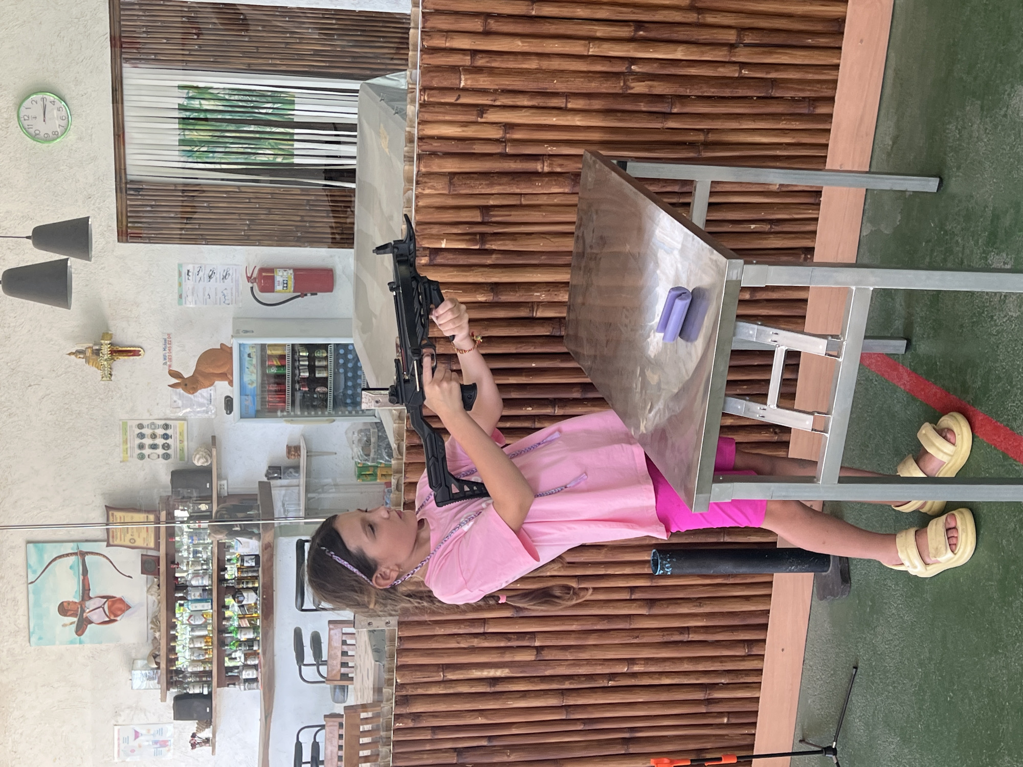 A young girl in a pink shirt and pink shorts is holding a black gun and aiming it while lying on a metal table. The scene appears to be an indoor shooting range in Baan Mae Nam, Thailand, judging by other items and signage in the room. The room also contains shelves with items displayed.