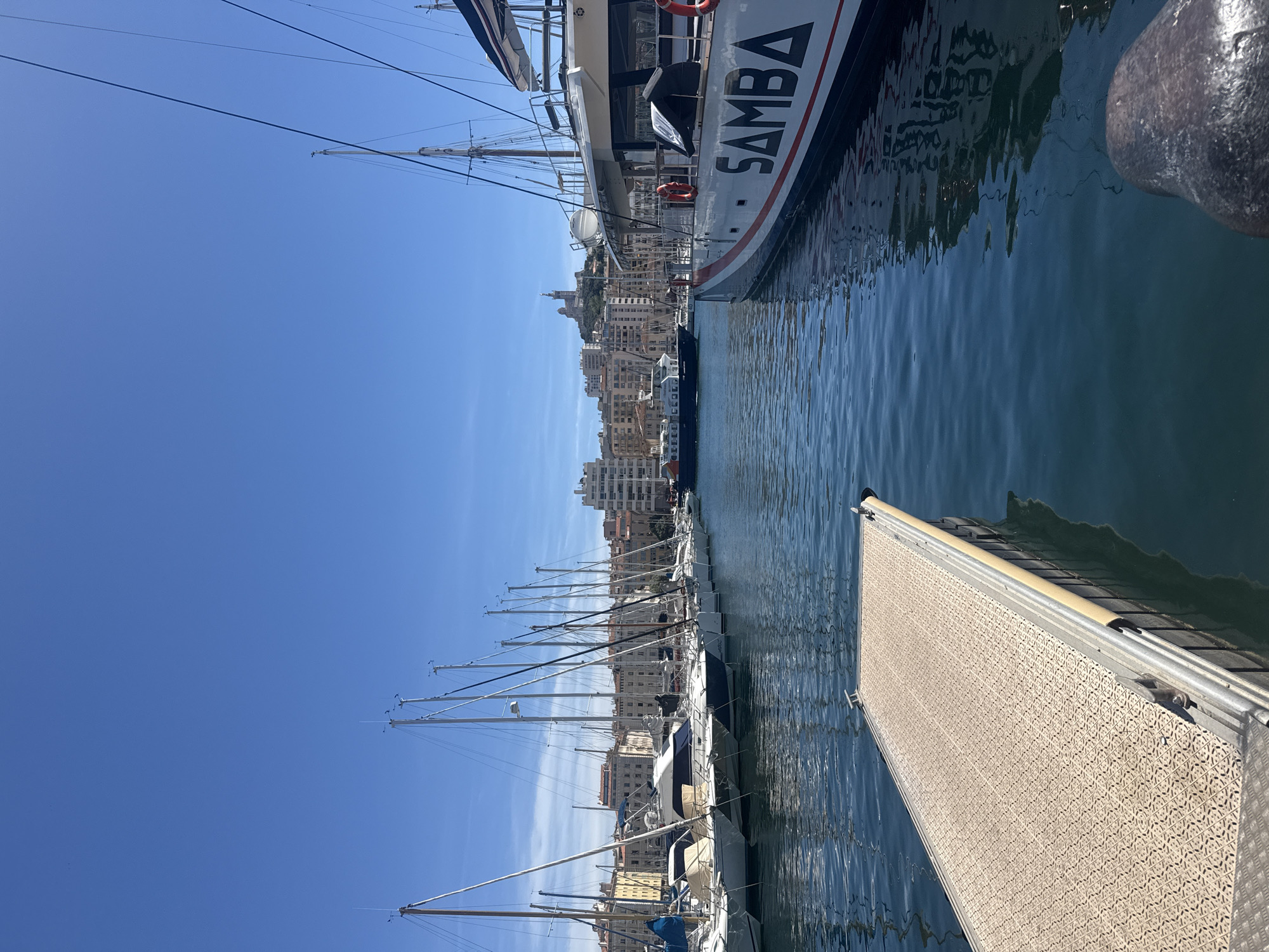 The image depicts a harbor scene under a clear blue sky. In the foreground, a textured, light-colored pontoon extends into the calm, blue water. To the right, a large white boat with red and black trim is docked, displaying the word "SAMBA" in large black lettering on its side. Life preservers are visible on the boat. In the middle ground, a line of sailboats with tall masts are moored, their white sails furled. Behind the sailboats, a cityscape with buildings of varying heights is visible, and on a hill in the distance, a prominent structure, possibly a church or monument, stands against the sky. A ferry or larger boat is navigating the water between the moored sailboats and the cityscape. The water reflects the clear sky and the surrounding structures. The overall impression is a sunny day at a bustling harbor.