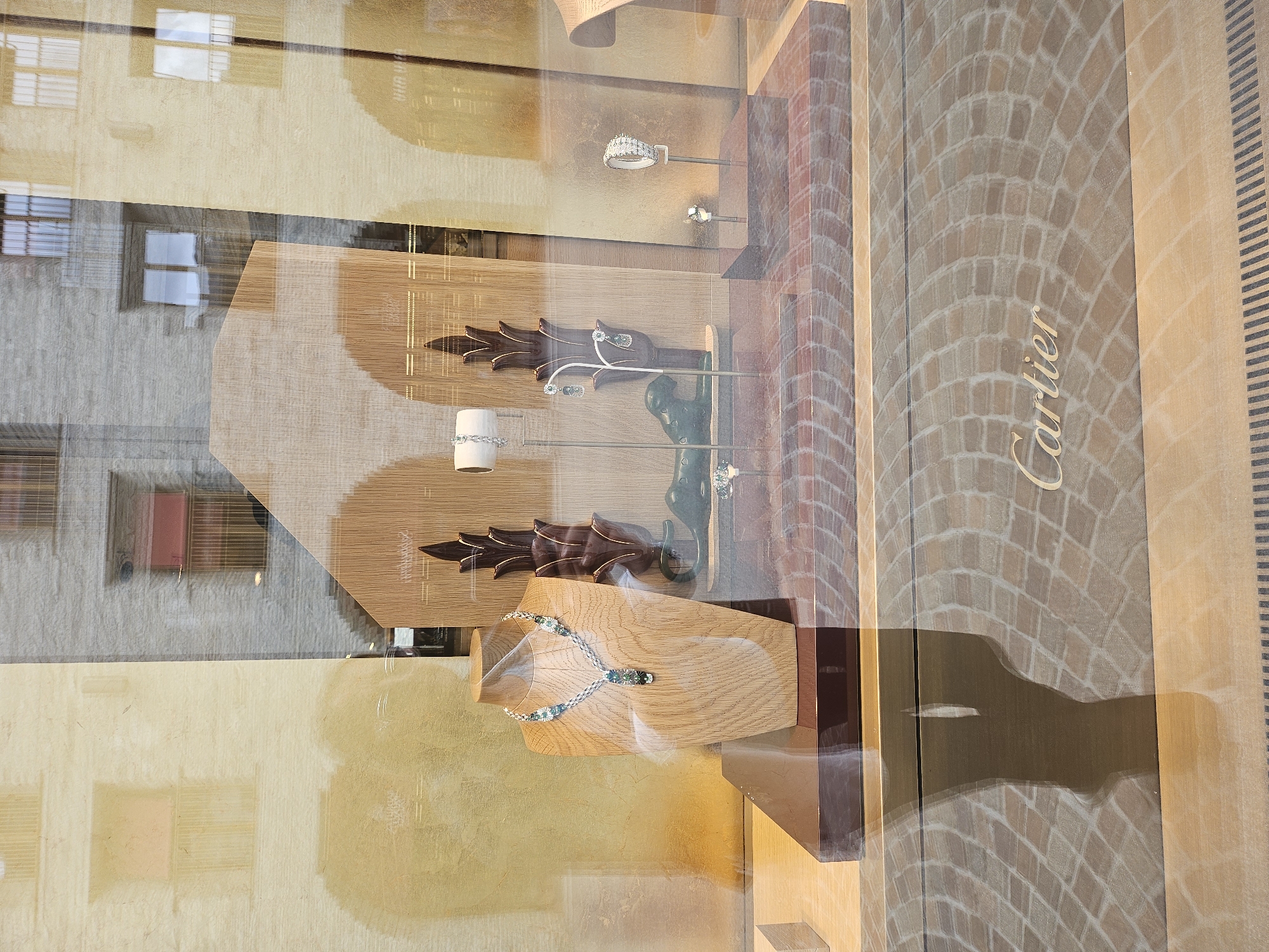 The image shows a shop window display, likely a jewelry store given the items on display. Inside the window, there are several pieces of jewelry, including rings and necklaces, presented on wooden stands. One prominent display features a necklace with blue and clear stones on a light-colored wooden bust. The background of the display has a brick pattern and a visible "Cartier" logo, indicating the brand of the store, located in St. Moritz, Switzerland. Reflections of the exterior, including buildings with windows, are visible on the glass of the shop window. The lighting suggests it is daytime, possibly late afternoon, with sunlight casting shadows. The scene appears calm and static, typical of a retail environment.