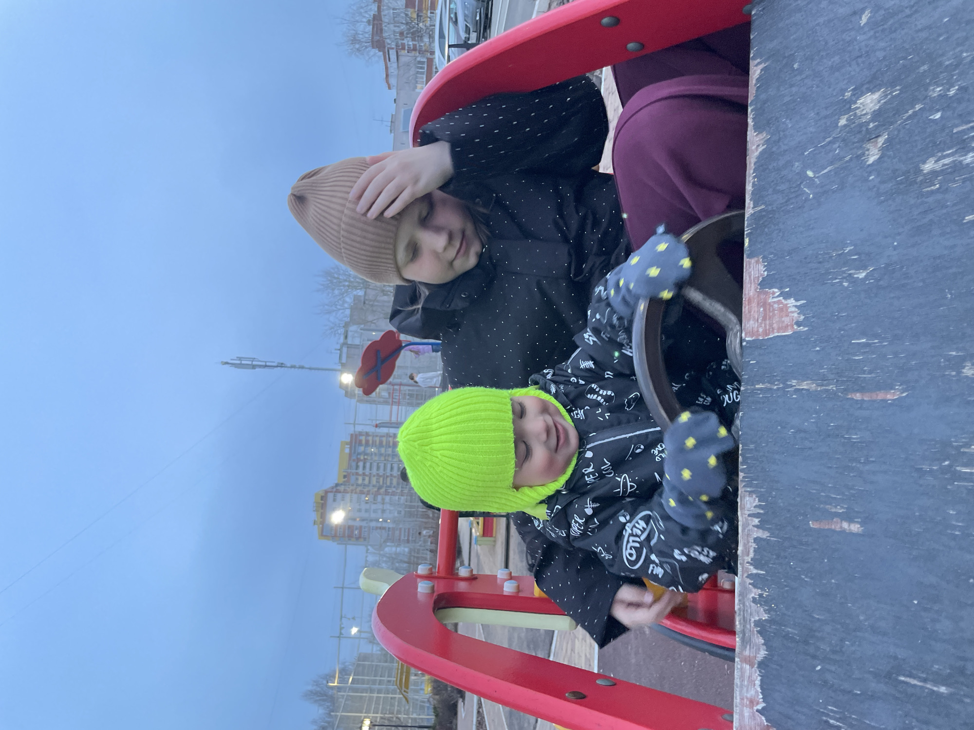 Two children are playing on a playground in Izhevsk, Russia. The older child wears a tan beanie, a black jacket with white polka dots, and maroon pants. She has her hand on her forehead. The younger child wears a neon green beanie, a black jacket with white graffiti print, and gloves with yellow polka dots. He is sitting and holding a steering wheel. The playground has red and white features. In the background are apartment buildings and streetlights. The sky is overcast.