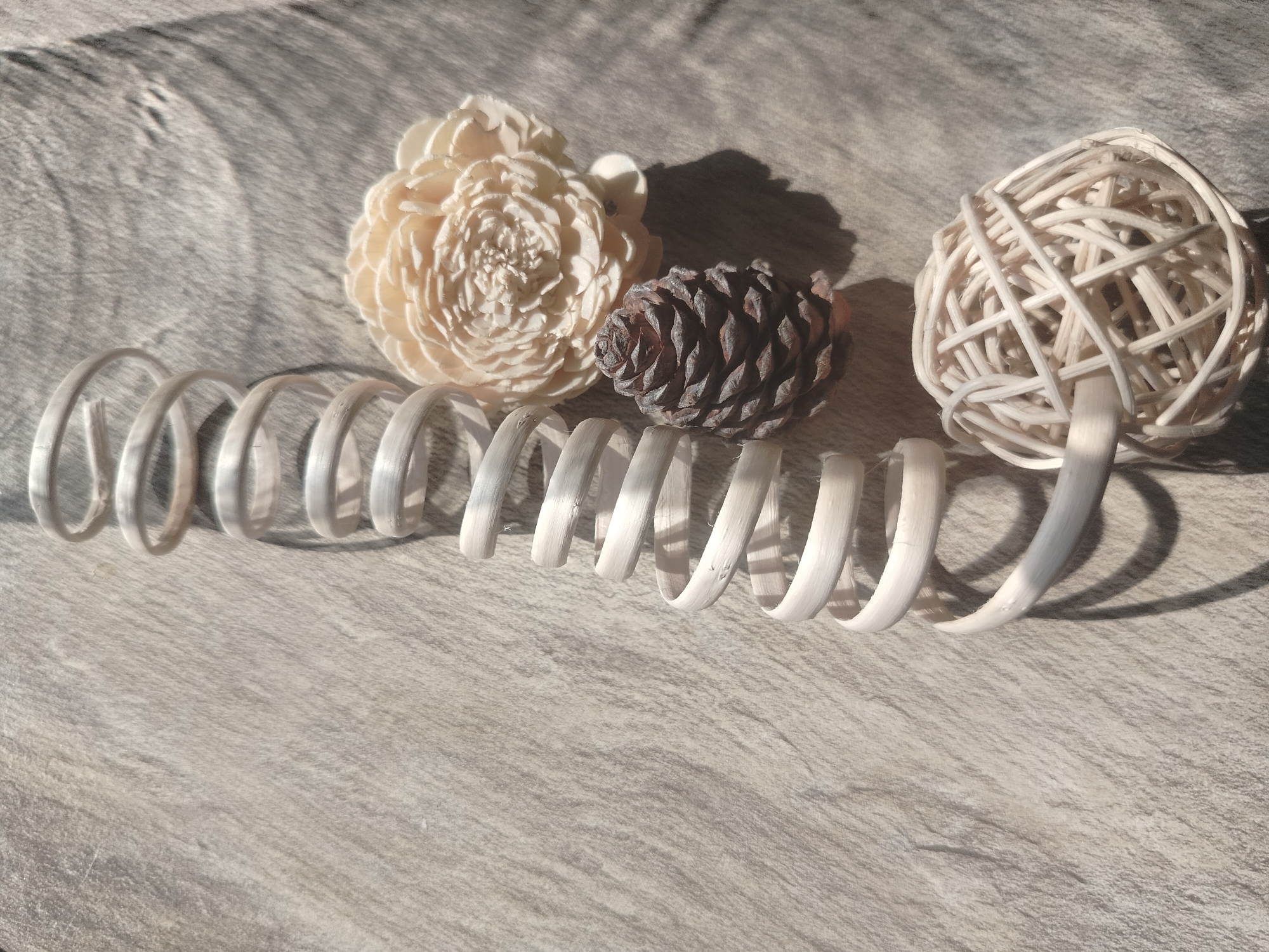 The image depicts a still life arrangement of decorative objects on a textured surface, likely a wooden table. The objects include a light-colored, layered decorative flower, a dark brown pine cone, and a white, open-weave sphere. To the left of these objects is a coiled, white decorative element that resembles a spring. The lighting suggests natural sunlight, casting shadows on the surface, indicating an indoor setting. There are no people or actions visible in the image. The location in Yashkino, Russia is not discernible from the visual information provided.