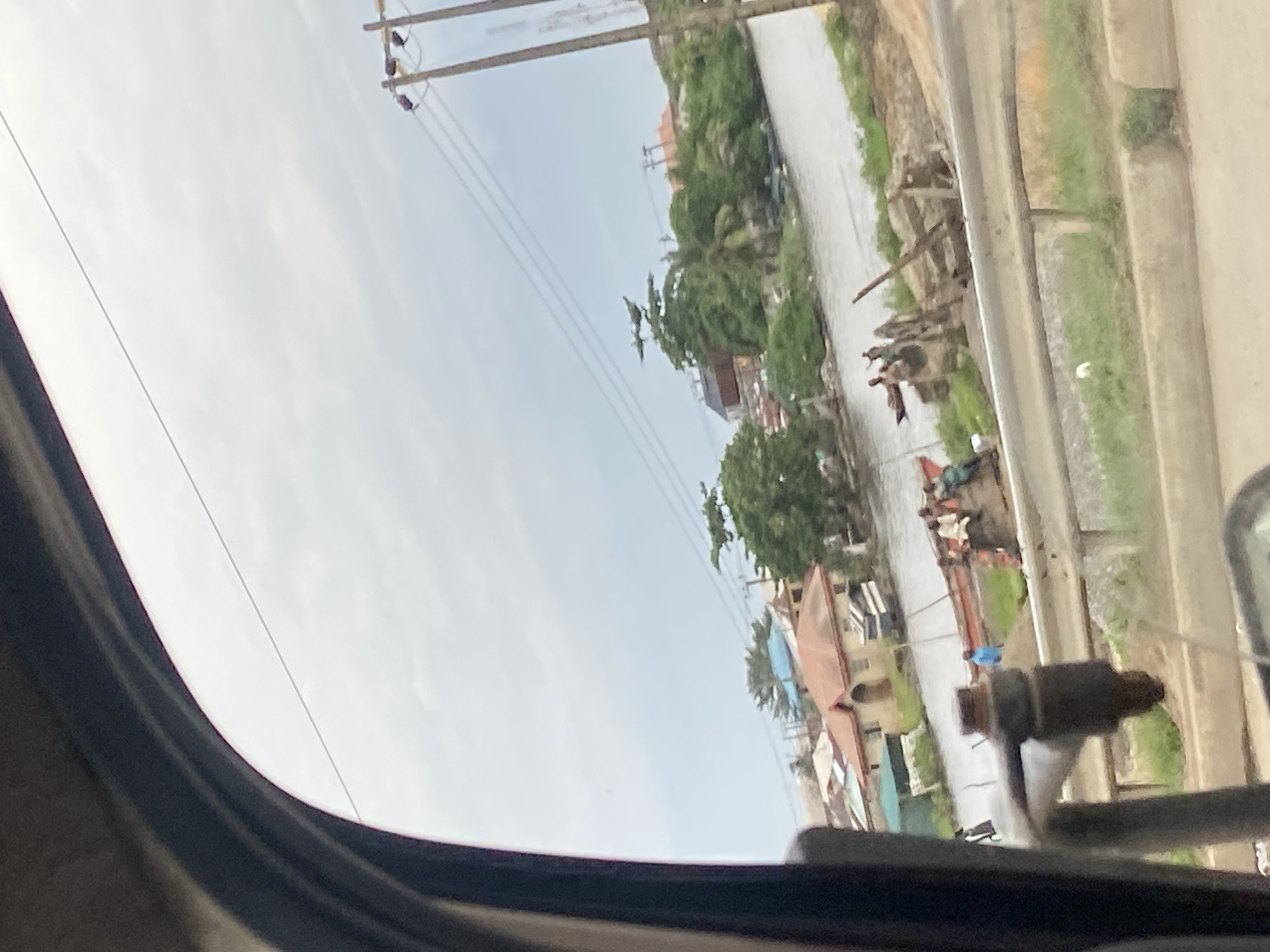 The image shows a tilted view of a street scene in Ikorodu, Nigeria, as seen from inside a vehicle. The sky is overcast, suggesting a cloudy day. A narrow body of water, possibly a canal or river, runs alongside the street, with green vegetation and some debris on its banks.
Several buildings, including houses and shops, are visible along the opposite side of the water. Some of these buildings have pitched roofs. Trees, some with dense foliage, are scattered throughout the scene. Power lines stretch across the sky, supported by utility poles, indicating the presence of electricity infrastructure.
In the foreground, the dark interior of the vehicle is visible, including a part of the dashboard and a metal fixture, likely a rearview mirror assembly. The perspective is from the passenger side of the vehicle, looking out.
Further down the street, a group of people are gathered near the water's edge. Some appear to be standing or sitting, and their activities are unclear due to the distance and the image's orientation. There are also what appear to be small boats or rafts on the water. A small blue object is visible near the water. The overall impression is of a bustling, possibly residential or commercial area situated near a waterway. No text is visible in the image.