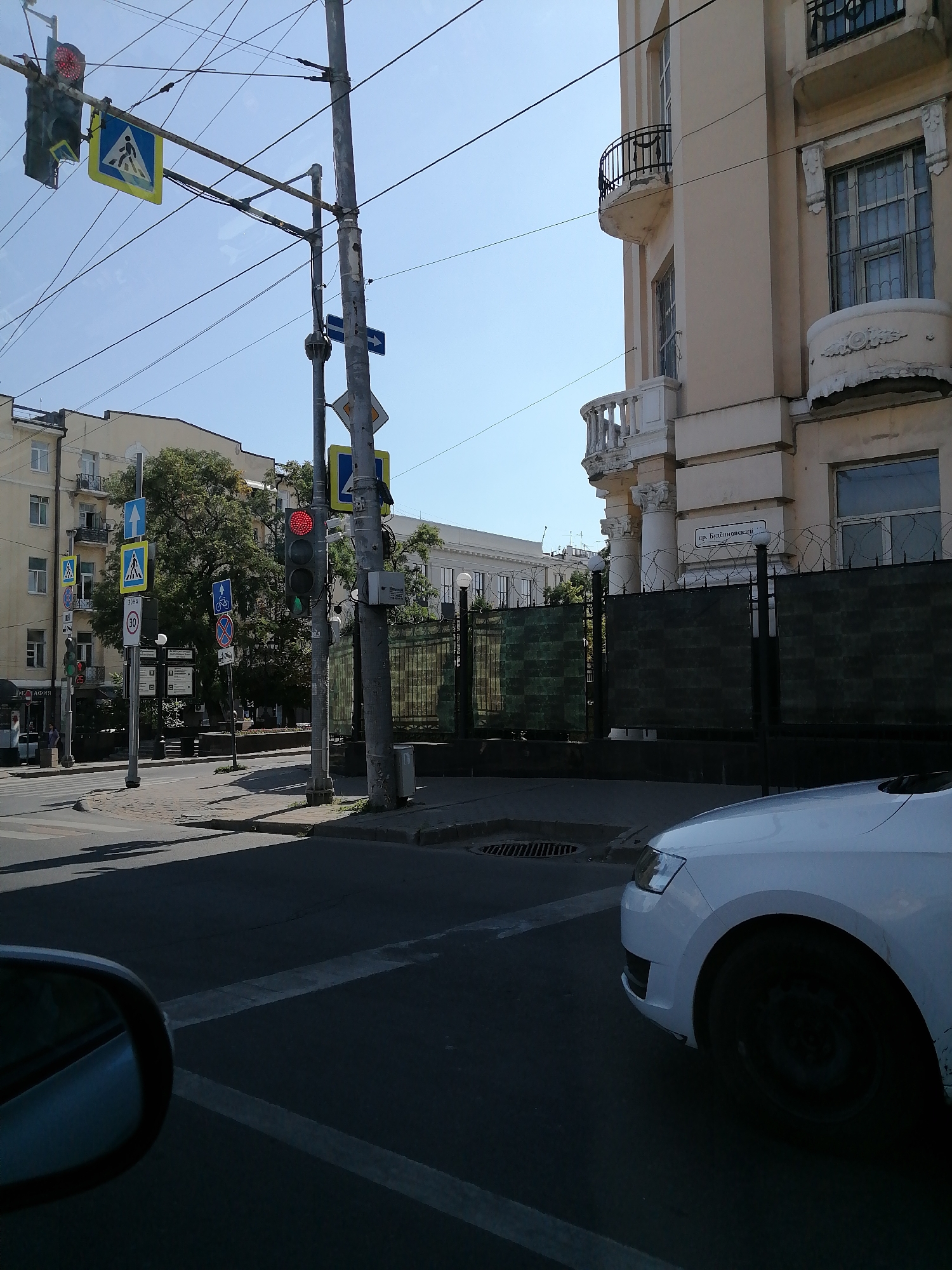 This is a street-level, daytime, medium shot captured from a vehicle in Rostov-on-Don, Russia. The image focuses on a street intersection with traffic lights and various road signs. A red traffic light is illuminated. A pedestrian crossing sign is visible. Building facades line the street. A white car is partially visible in the foreground, and a car's side mirror is also in view. The sky is clear and blue. Overhead wires crisscross the scene. A metal fence with barbed wire adorns the top is also visible. A street sign with Cyrillic lettering is mounted on a pole.