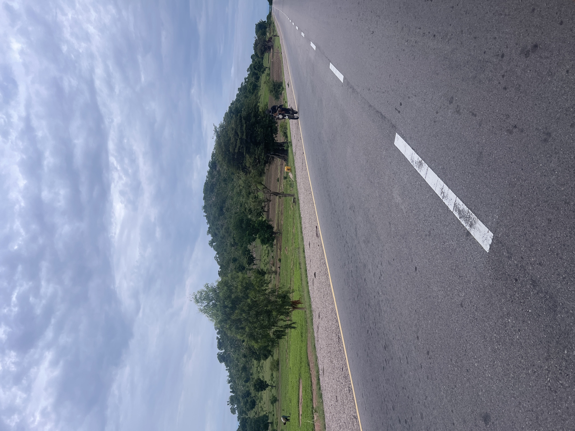 In the city of Minda, Nigeria, a paved road stretches into the distance under a cloudy sky. The road is marked with white dashed lines. On the left side of the road, a green field slopes upwards towards a densely wooded hill. A single person is visible in the field, tending to the crops. A motorbike carrying one person rides down the road, heading away from the viewer. The general environment is rural, with natural vegetation and agricultural land bordering the road.