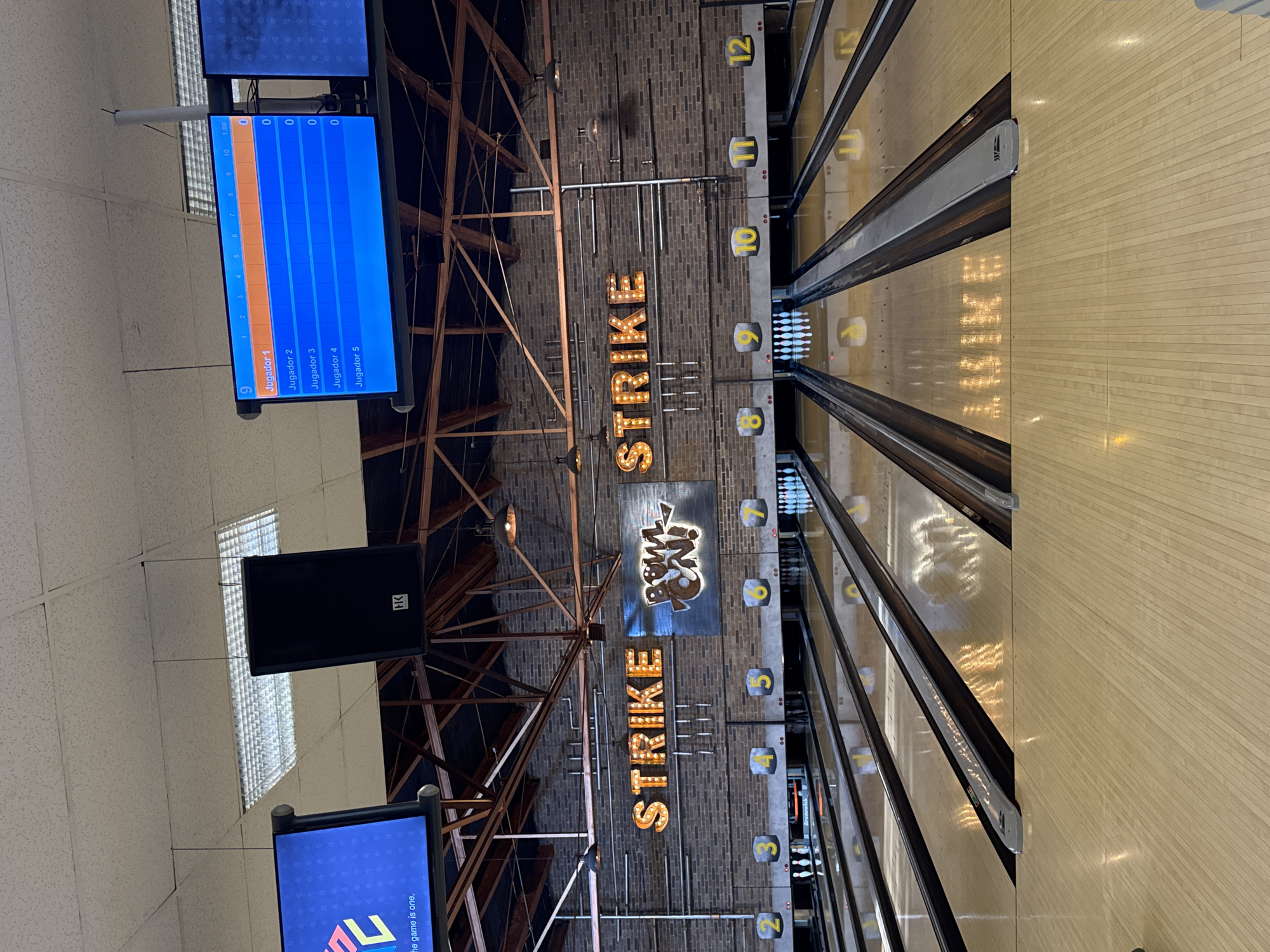 The image depicts the interior of a bowling alley. There are multiple bowling lanes visible, with pins set up at the end of several lanes. Above the lanes, there's brickwork adorned with "STRIKE" signs and numbered markers indicating lane numbers (2 through 12). Scoreboards displaying player names and scores are present, one displaying "Jugador 1" through "Jugador 5." The image includes ceiling structures with lights and a "Bowl" logo. Based on the OCR, the image might contain a screen displaying "El game is one." The bowling alley appears to be located in the city of Puebla, Mexico.