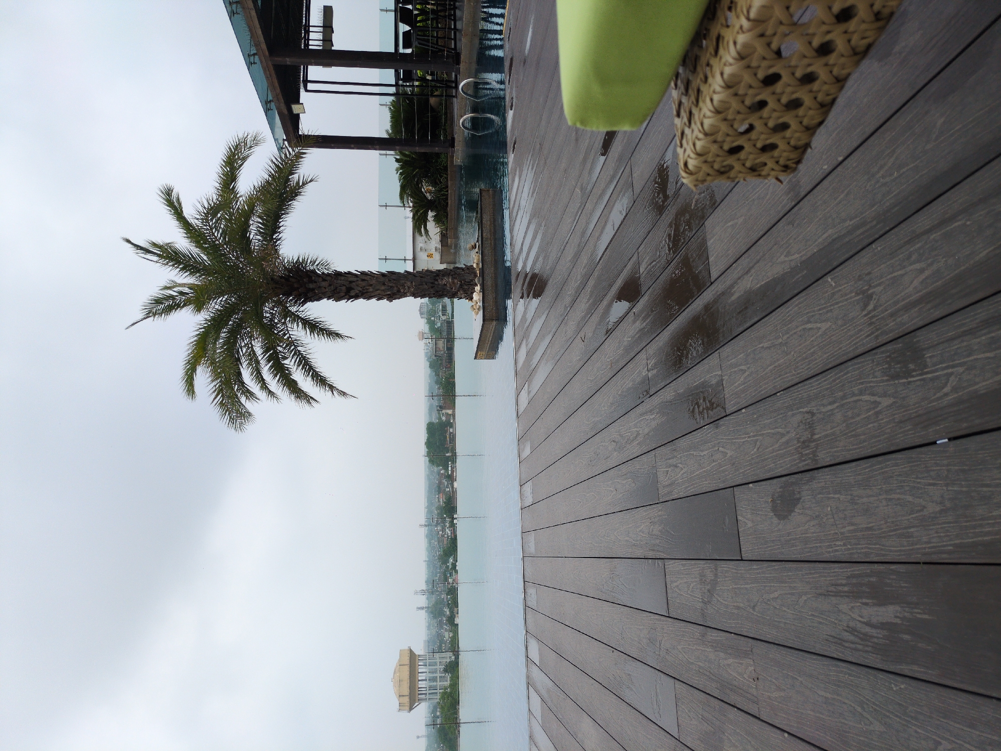 The image captures a serene poolside scene, likely at a rooftop establishment in Agra, India, given the presence of a distinctive water tower in the background that is characteristic of the city. In the foreground, a wooden deck, wet with scattered puddles, stretches out, its dark planks laid horizontally. To the right, a corner of a wicker ottoman with a bright green cushion is visible, suggesting a seating area.
Dominating the mid-ground is an infinity pool, its surface reflecting the overcast sky. A solitary palm tree stands within the pool, its trunk surrounded by a small stone planter. Beyond the pool, a cityscape unfolds under a hazy sky, with buildings and telecommunication towers dotting the horizon. A prominent, pale yellow water tower, a landmark in Agra, is clearly visible to the left, confirming the location. To the right of the palm tree, a covered seating area with wooden pillars and a glass roof can be seen, furnished with bar stools. The overall atmosphere is tranquil, hinting at a peaceful retreat, despite the cloudy weather. There are no people visible in the frame, focusing the viewer's attention on the architectural elements and the expansive view.
