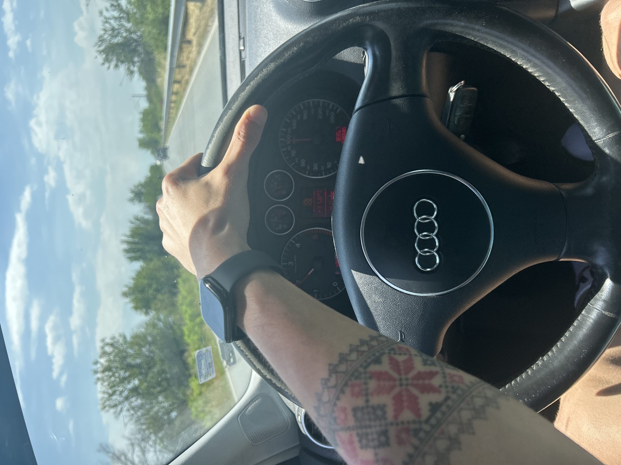 The image captures a first-person perspective from inside a car, looking out through the windshield and towards the dashboard. The primary subject is the driver's left arm and hand gripping the steering wheel. The driver is wearing a dark smartwatch on their left wrist, and their forearm is adorned with a colorful tattoo featuring a traditional geometric pattern in red and black.
The scene depicts the activity of driving. The steering wheel, bearing the Audi logo, dominates the central-right portion of the frame. Behind it, the car's instrument cluster is visible, with the speedometer needle indicating approximately 60-70 km/h. The odometer clearly displays "235606" km, and the engine temperature gauge shows roughly 90 degrees Celsius. A car key is partially visible tucked near the steering column.
Through the windshield, a road with a guardrail runs parallel to the car, indicating travel on a highway or main road. Trees and green foliage line the side of the road. The sky is bright with scattered white clouds, suggesting a sunny daytime setting. The image itself is rotated 90 degrees counter-clockwise. The location context indicates this scene takes place in Chernozemen, Bulgaria. There is a sticker on the windshield, but its text is illegible.