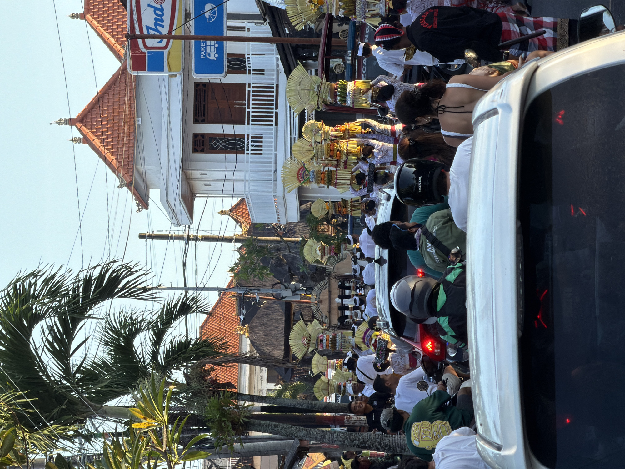 In Mengwi, Indonesia, a street scene unfolds with a Balinese Hindu procession in progress. People in traditional white clothing and headdresses carry ornate, tiered offerings decorated with yellow fans and fruits. The procession is flanked by onlookers and a stalled traffic line of cars and motorcycles. Buildings with red-tiled roofs and signs featuring the word "Indo" are visible in the background. Palm trees line the street.