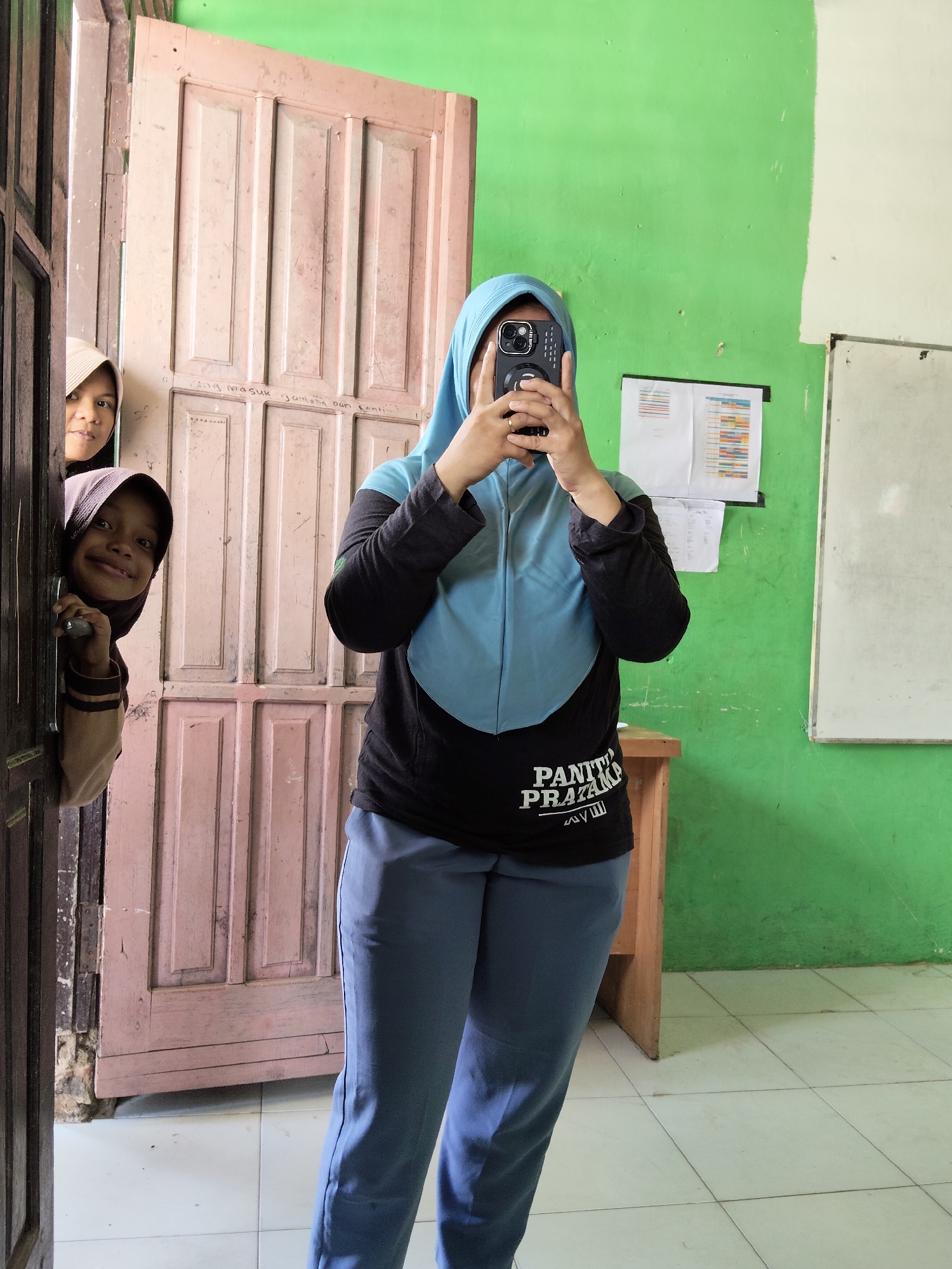 In a classroom in Lori, Indonesia, two young girls wearing hijabs peek out from behind a pink wooden door. One girl, with a dark brown hijab, is leaning out and smiling directly at the camera. The other girl, wearing a lighter colored hijab, is partially visible behind the first. A young woman, also wearing a hijab and a black shirt with a light blue overlay, stands in the center of the frame, holding a smartphone to take a selfie. She is wearing light blue pants. The classroom walls are painted bright green and white. A wooden desk and a whiteboard are visible in the background. The floor is tiled with white squares. The scene appears to be a candid moment of children interacting with the photographer in a school environment.
