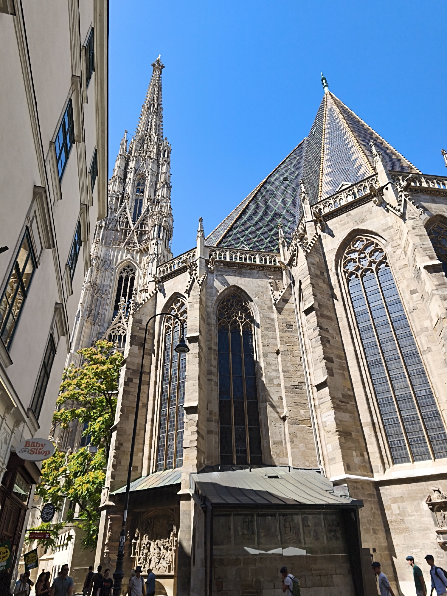 The image shows a street view in Vienna, Austria, featuring the St. Stephen's Cathedral. The cathedral's tall, ornate spire and tiled roof are prominent in the frame, set against a clear blue sky. To the left, the side of a lighter-colored building with arched windows runs along the street. Several people are walking along the cobblestone street, some are looking at the cathedral, while others are walking by. A street lamp with a single light fixture is visible in the middle of the image. Signs for businesses, including "Baby Shop" and "Fankl Kaffee," are attached to the buildings. The architecture is predominantly Gothic, with intricate stone carvings and tall, slender windows. The scene is bathed in sunlight, casting shadows on the street and buildings.
