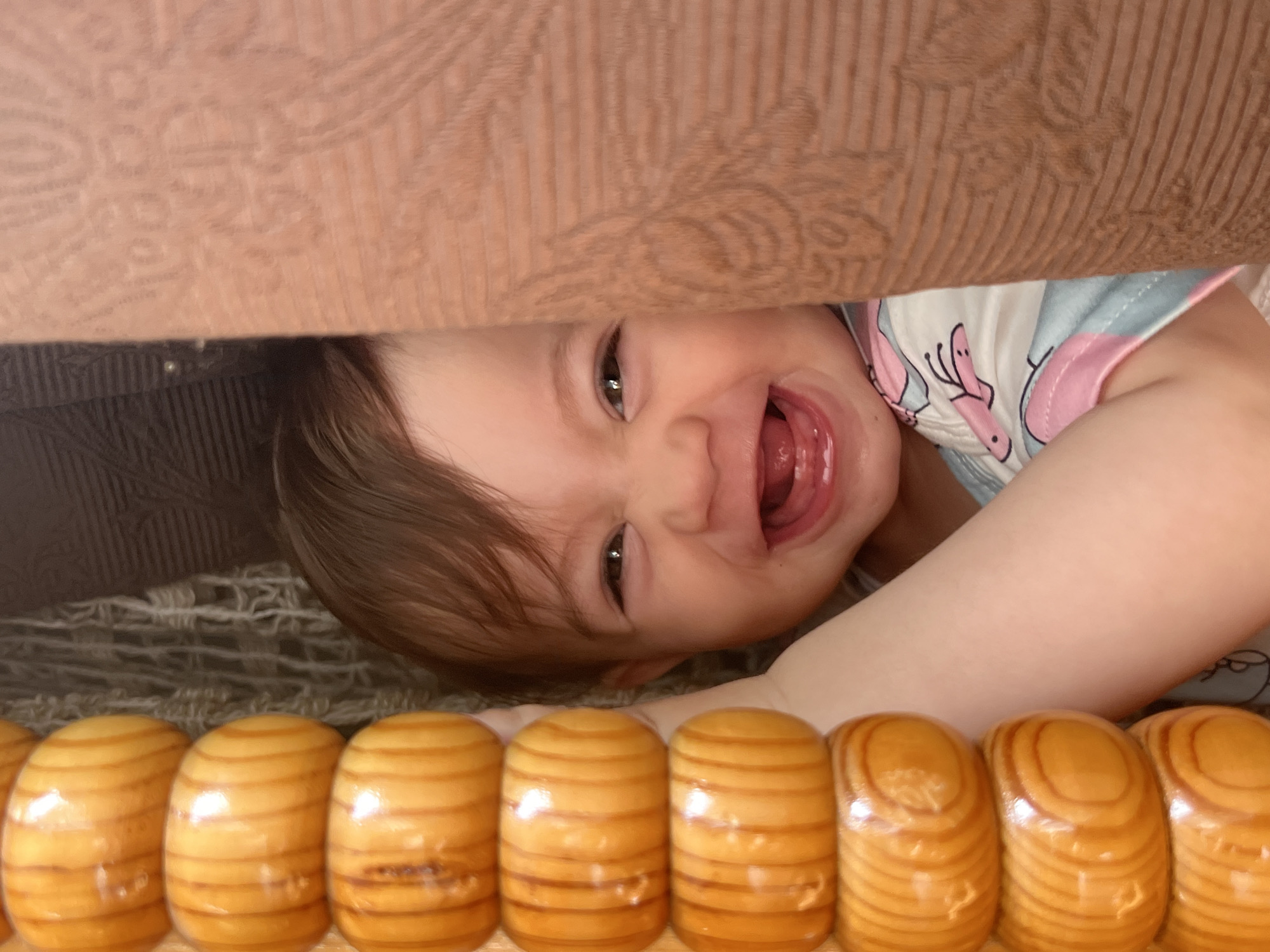 A close-up, angled shot captures a joyful baby peeking out from underneath what appears to be a piece of furniture. The baby's face is turned sideways, with bright brown eyes looking directly at the camera. A wide, open-mouthed smile reveals their tongue and a few developing teeth, conveying an expression of clear happiness and playful laughter. Their light brown hair frames their face.
Above the baby's head, a textured, light beige or tan fabric is visible, likely upholstery from a couch or bed, featuring a subtle, embossed pattern. This fabric forms the upper boundary of the narrow space from which the baby is emerging. Below the baby's head, a dark, somewhat textured fabric or surface is partially visible.
In the foreground, running along the bottom edge of the image, is a row of shiny, light brown wooden beads or elongated segments. These objects display natural wood grain lines and appear to be part of a wooden toy, a decorative element, or a barrier, possibly resembling large abacus beads or a segment of a playpen railing.
The scene suggests an indoor home setting, likely during the daytime, with the baby engaged in a moment of playful interaction. There are no specific cues within the image itself to indicate the provided location of Elena, Bulgaria, nor is any text visible in the photograph.