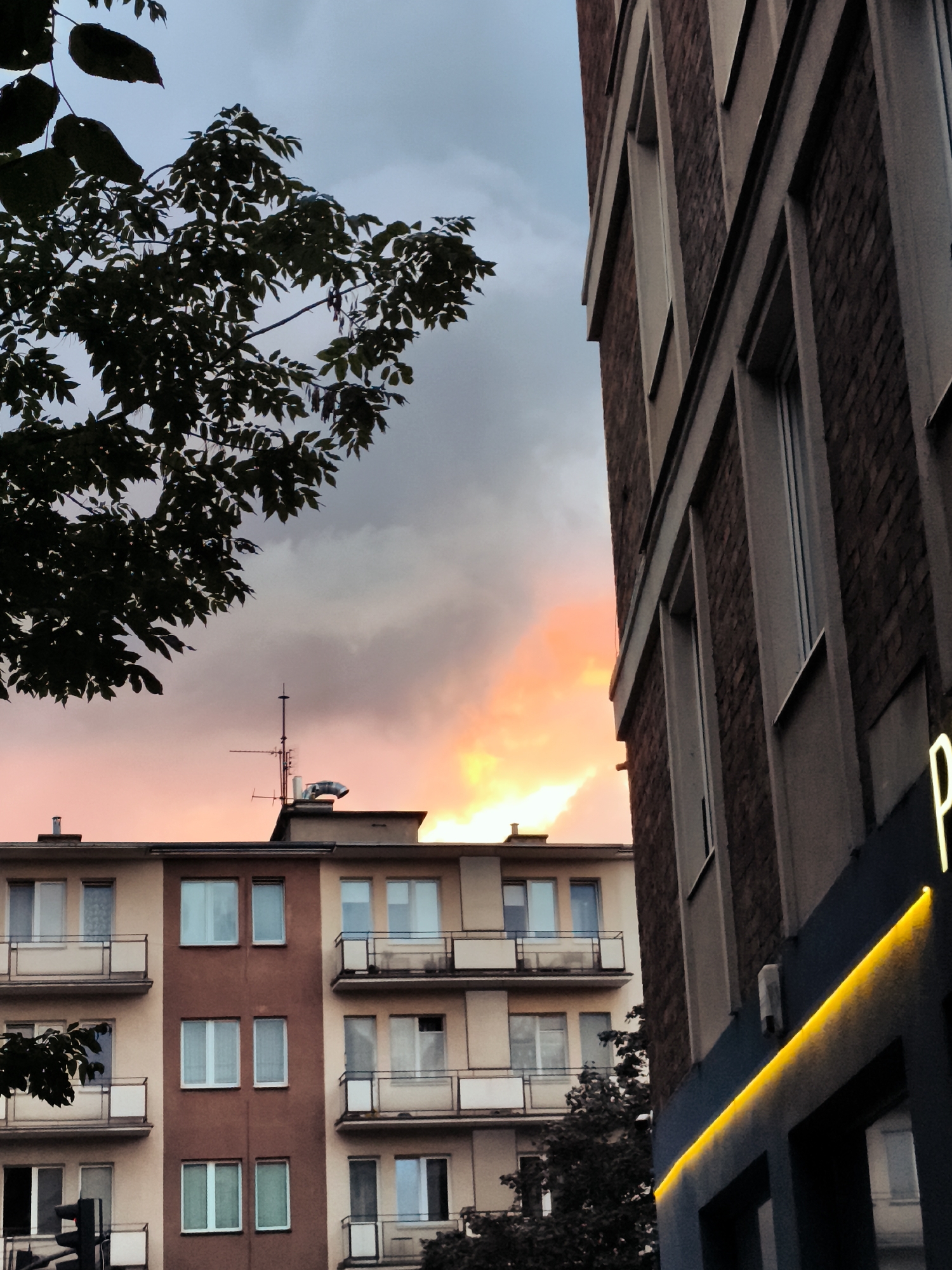 The image depicts a street view in Gdańsk, Poland, featuring an apartment building and a brick building under a dramatic sky. The apartment building, situated on the left, has a light-colored facade with multiple windows and balconies. The brick building on the right has a darker facade and a prominent neon light that casts a yellow glow. The sky above is filled with dark clouds, with a bright orange and yellow hue illuminating the horizon, suggesting either sunrise or sunset. A tree with dark leaves is visible in the foreground on the left, partially obscuring the view of the apartment building. There are no people visible in the image.