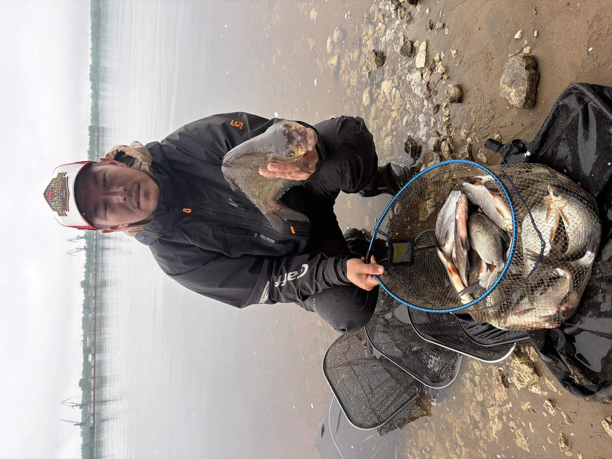 An angler, wearing a dark jacket with a light-colored hood and a red and white cap, crouches by the water's edge, holding a large fish in his hands. The fish appears to be a bream or similar species, with metallic scales. To his right, a large fishing net (keepnet) filled with numerous other fish, likely a mix of different types, rests on the muddy, rocky shore, partially submerged in shallow water.
The man is looking towards the camera with a neutral expression. The cap has the text "MAXIMUS" prominently displayed. His jacket sleeve shows the brand "Craft" and other smaller text. The setting is an outdoor body of water, possibly a large river or lake, with a wide, calm surface reflecting the overcast sky. On the distant shore across the water, industrial-looking structures, possibly cranes, and a line of trees are faintly visible through the hazy conditions. The immediate shoreline is a mix of wet sand and various sizes of rocks and pebbles. The water near the shore is shallow and somewhat murky. The overall atmosphere suggests a cool, cloudy day, typical for fishing. The image captures a successful fishing outing in what appears to be Noviy, Russia.