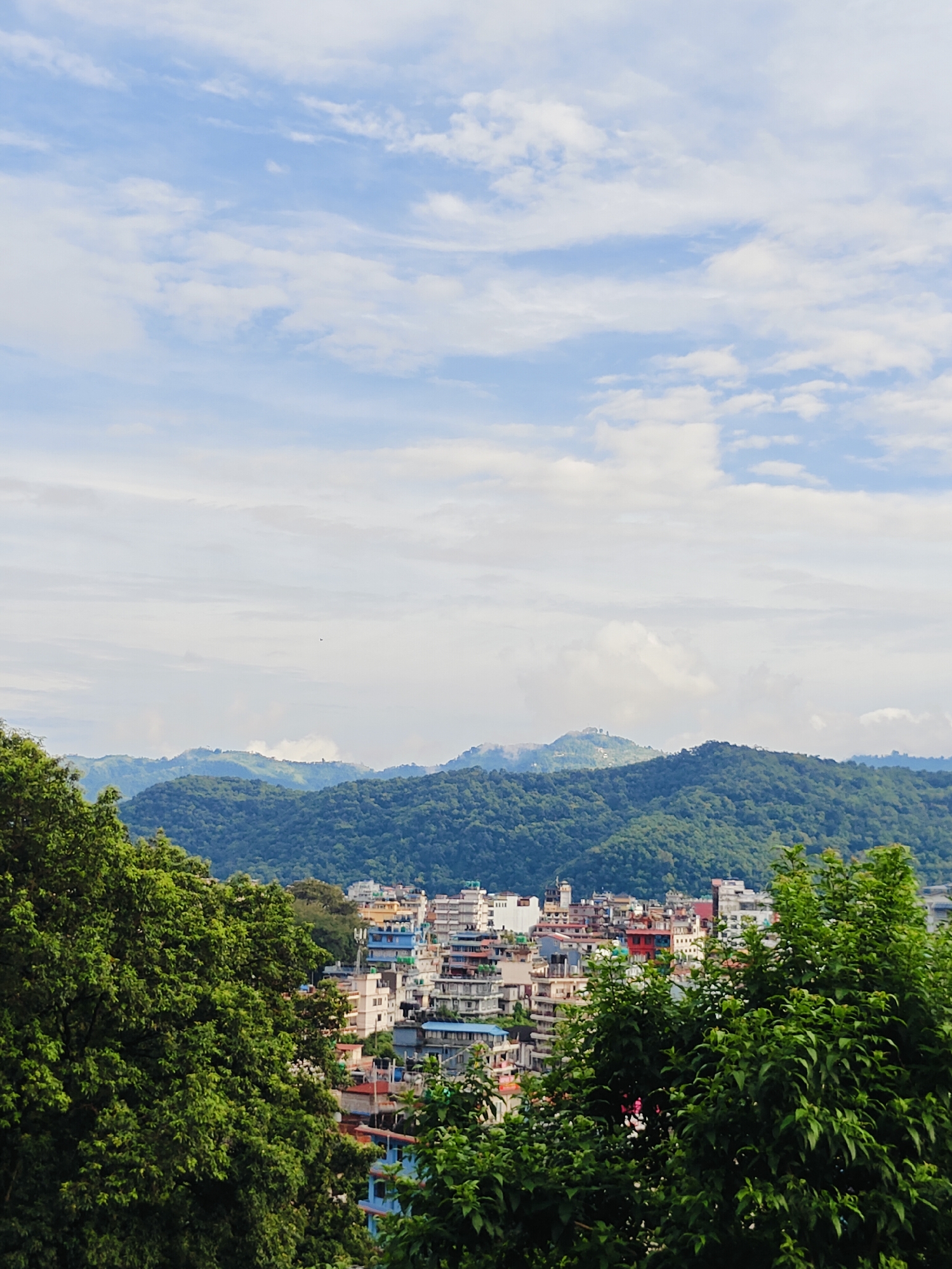 The image shows a panoramic view of the city of Pokhara, Nepal, as seen from a slightly elevated vantage point. In the foreground, lush green trees, with their dense foliage, partially obscure the view of the city. The city itself is characterized by a cluster of multi-story buildings, displaying a variety of architectural styles and colors, ranging from white and beige to blue and red. These buildings are densely packed, indicating an urban environment.
Beyond the city, a series of rolling hills covered in dense green vegetation rise up, forming a verdant backdrop. These hills appear to be part of the larger Annapurna mountain range, a prominent geographical feature of the region. The sky above is a light blue, dotted with scattered white clouds, suggesting a partly cloudy day. The overall impression is one of a city nestled within a beautiful natural landscape, showcasing the harmonious coexistence of urban development and the surrounding natural environment. No people are visible in the image.