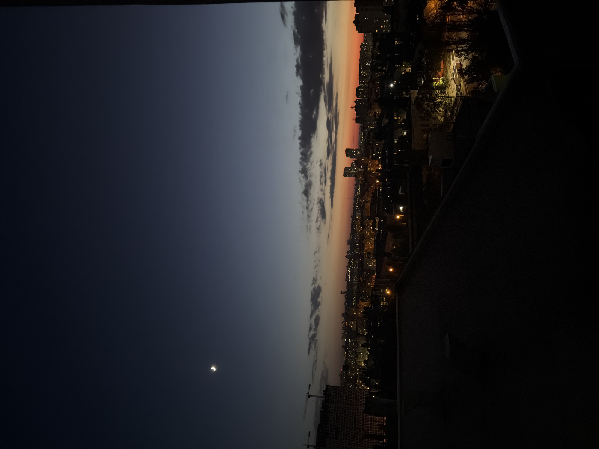This image captures a twilight cityscape of Kyiv, Ukraine, from a high vantage point, likely a rooftop. The sky transitions from a deep navy at the top to a soft orange and red hue near the horizon, indicating either sunrise or sunset. A crescent moon is visible in the upper left quadrant of the sky, accompanied by a faint star or planet.
The city below is illuminated by a scattering of lights from buildings and streetlamps, suggesting it is dusk or night. Several multi-story buildings, some appearing to be under construction with cranes visible, dominate the left side of the frame. In the mid-ground, a cluster of taller buildings forms a distinct skyline. The foreground is occupied by the dark, silhouetted roof of the viewer's location, obscuring details of any immediate surroundings. Trees and other urban foliage are visible scattered throughout the city, with some illuminated by streetlights. The overall impression is a serene yet vibrant urban panorama as day transitions into night in the Ukrainian capital.