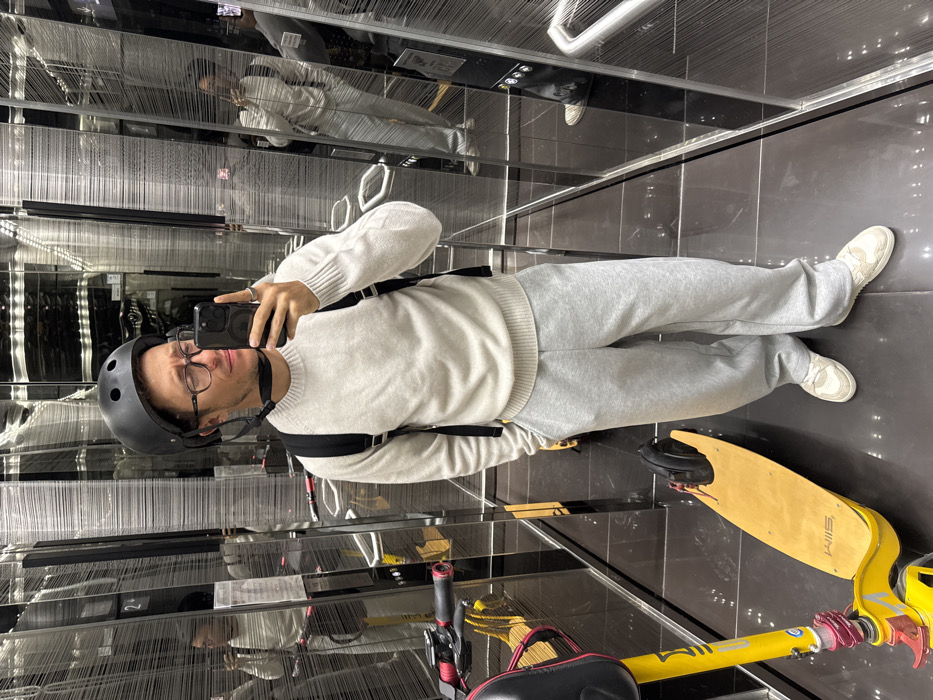 An adult male is depicted taking a selfie inside a modern elevator located in Tel Aviv-Yafo, Israel. The subject, appearing to be in his late 20s or early 30s, wears a black cycling helmet with a fastened chin strap, dark-framed eyeglasses, a light-colored long-sleeved crew-neck sweater, light grey sweatpants, and light-colored sneakers with white soles. He wears a black backpack over both shoulders and has a silver ring on his left ring finger. He holds a black smartphone in his right hand, pointed towards himself, and displays a slight squint or grimace. To his left, a yellow electric kick scooter with a wooden deck, bearing the 'SLIM' brand name, leans against the elevator wall. The scooter features black wheels, red accents on its frame and handlebars, and a black seat with a red underside. The elevator interior consists of dark, highly reflective walls, likely mirrored or polished metal, which create multiple reflections of the man and the scooter. The floor is dark and reflective. A silver handrail is partially visible in the reflections on the right side of the elevator car.