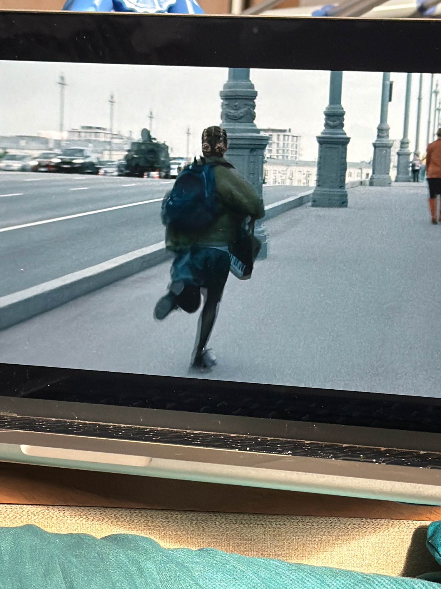 This is a medium-shot, eye-level, slightly blurry photograph of a person running across a bridge with a backpack. The person is seen from the back and is wearing a green jacket, dark shorts, and dark leggings. The person has a dark backpack on their shoulders. The bridge has decorative lampposts and a concrete barrier along the side. In the background, there are cars and buildings visible. The scene appears to be set in Khabarovsk, Russia, possibly on the Amur River bridge, a well-known landmark. The lighting suggests it might be daytime.