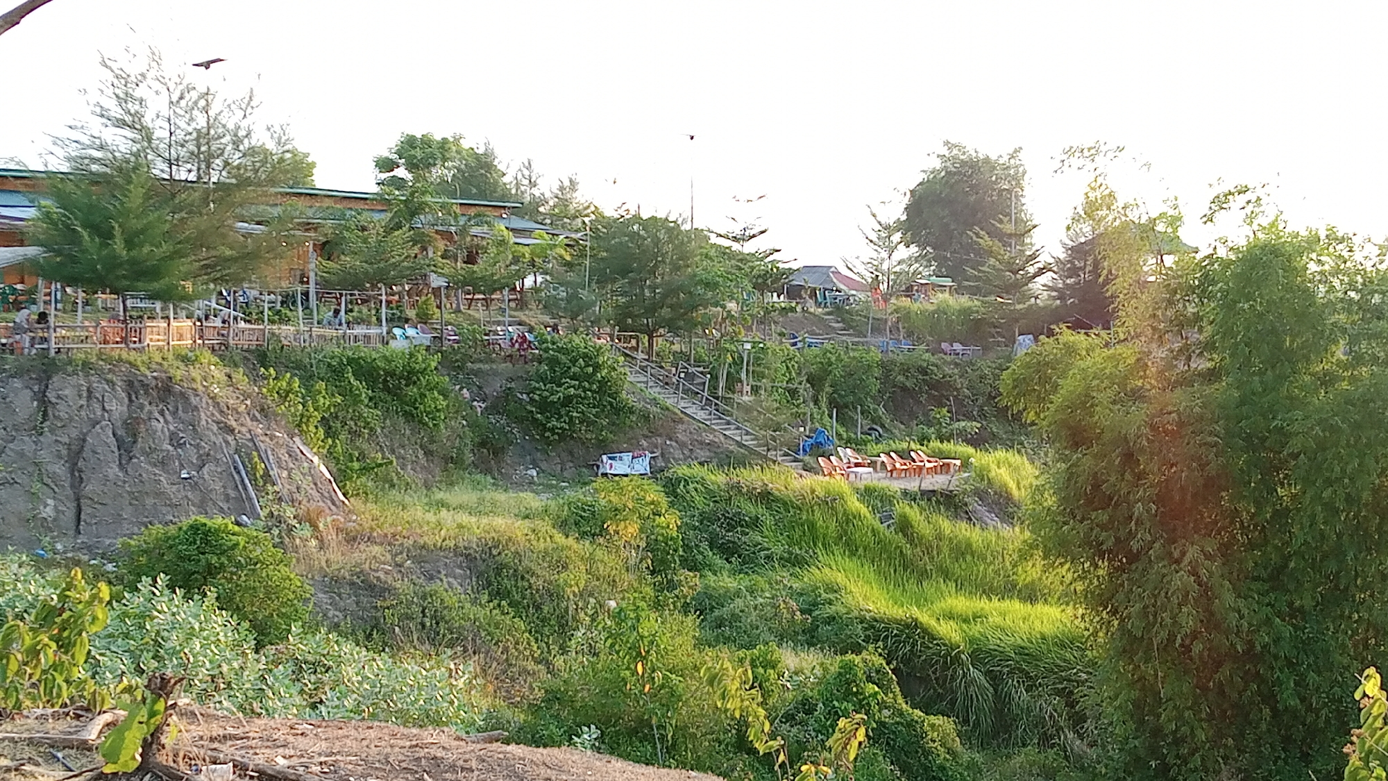 The image depicts an outdoor seating area and elevated structures overlooking a vegetated slope. The setting appears to be a park or restaurant, likely in Blangtingkeum, Indonesia, given the context. In the upper portion of the frame, several wooden tables and chairs are arranged on a raised platform, surrounded by trees and foliage. A long, low-slung building with a greenish roof is visible behind the seating area, suggesting it could be part of a restaurant or establishment. A flight of stairs descends the slope from the upper level, leading to a lower area with rows of orange chairs and tables. The slope itself is covered in lush green grass and various plants, with a rocky cliff face visible on the left side of the frame. A small, white cart or sign is placed near the base of the stairs. The overall scene is illuminated by natural light, suggesting it is daytime. There are a few indistinct figures seated at some of the tables in the upper area, and one person can be seen standing near the orange chairs on the lower level.