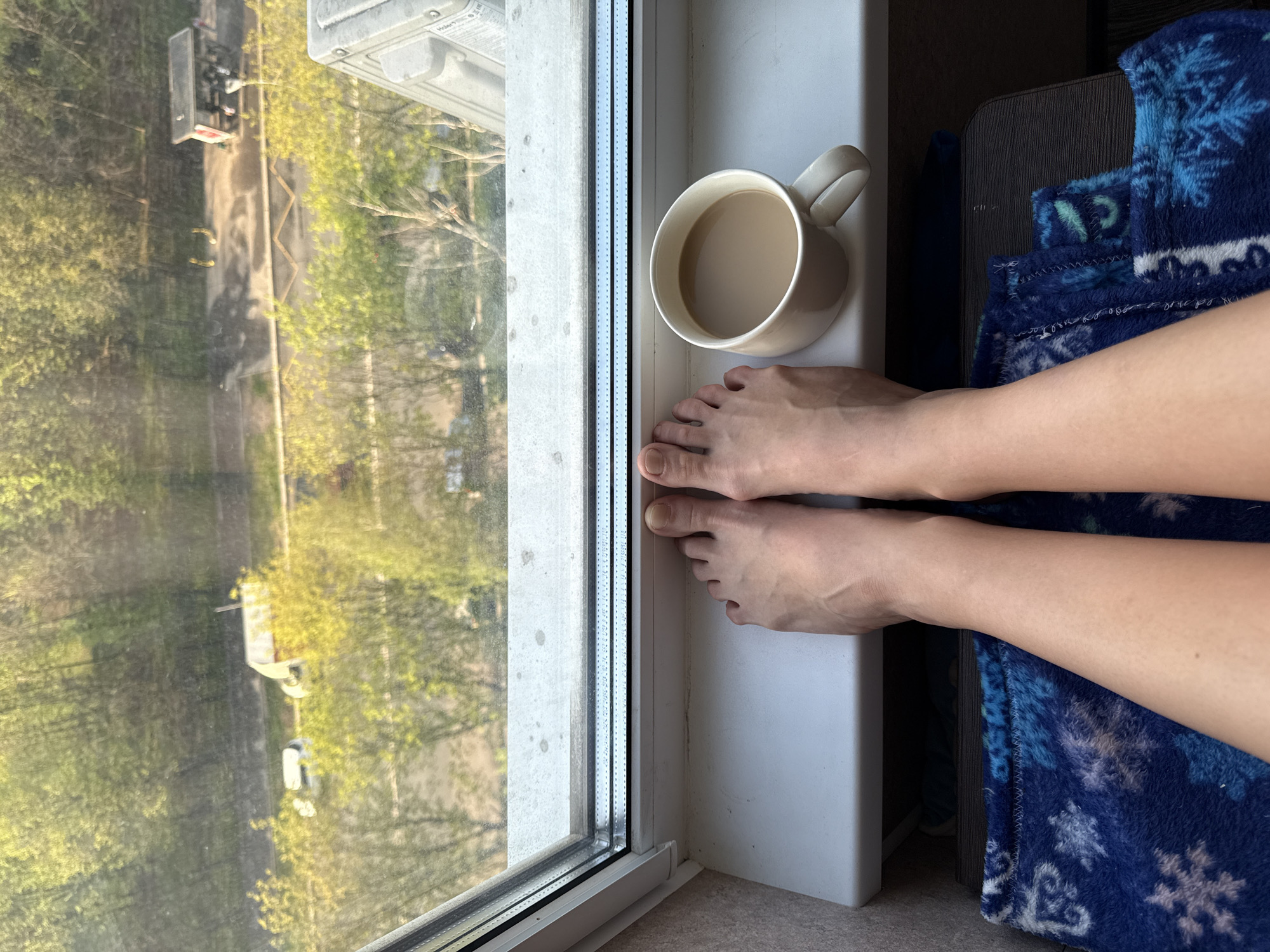 A high-angle interior shot of a window sill, featuring a coffee mug, human feet, and a glimpse of the outside world.
The window is located in a building, with a view overlooking a street in an Unknown, Unknown city. Green trees line the street, and a vehicle, possibly a truck or large utility vehicle, is visible.
The window sill is a white color and appears to be clean. A white ceramic coffee mug, filled with a light brown beverage, rests on the sill. Two bare human feet are positioned on the sill near the mug. The feet appear to be resting.
To the right of the feet is a blue blanket with white snowflake patterns. The blanket is draped over an unseen object.